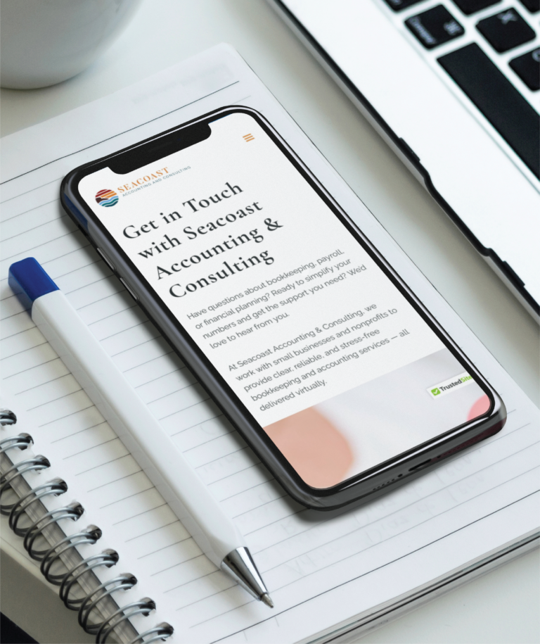 A smartphone displaying Seacoast Accounting & Consulting’s redesigned website rests on a spiral notebook next to a blue-and-white pen. The scene, set beside a laptop keyboard, highlights modern accounting support in New Hampshire on a clean white desk.