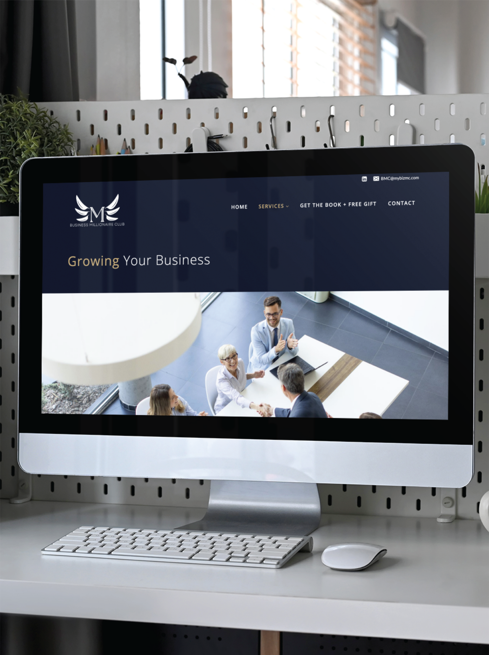 A computer screen displays a business website focused on educational consultants, featuring a header and a photo of professionals in a meeting. The desk has a white keyboard, mouse, and plant nearby, with natural light streaming through the window.