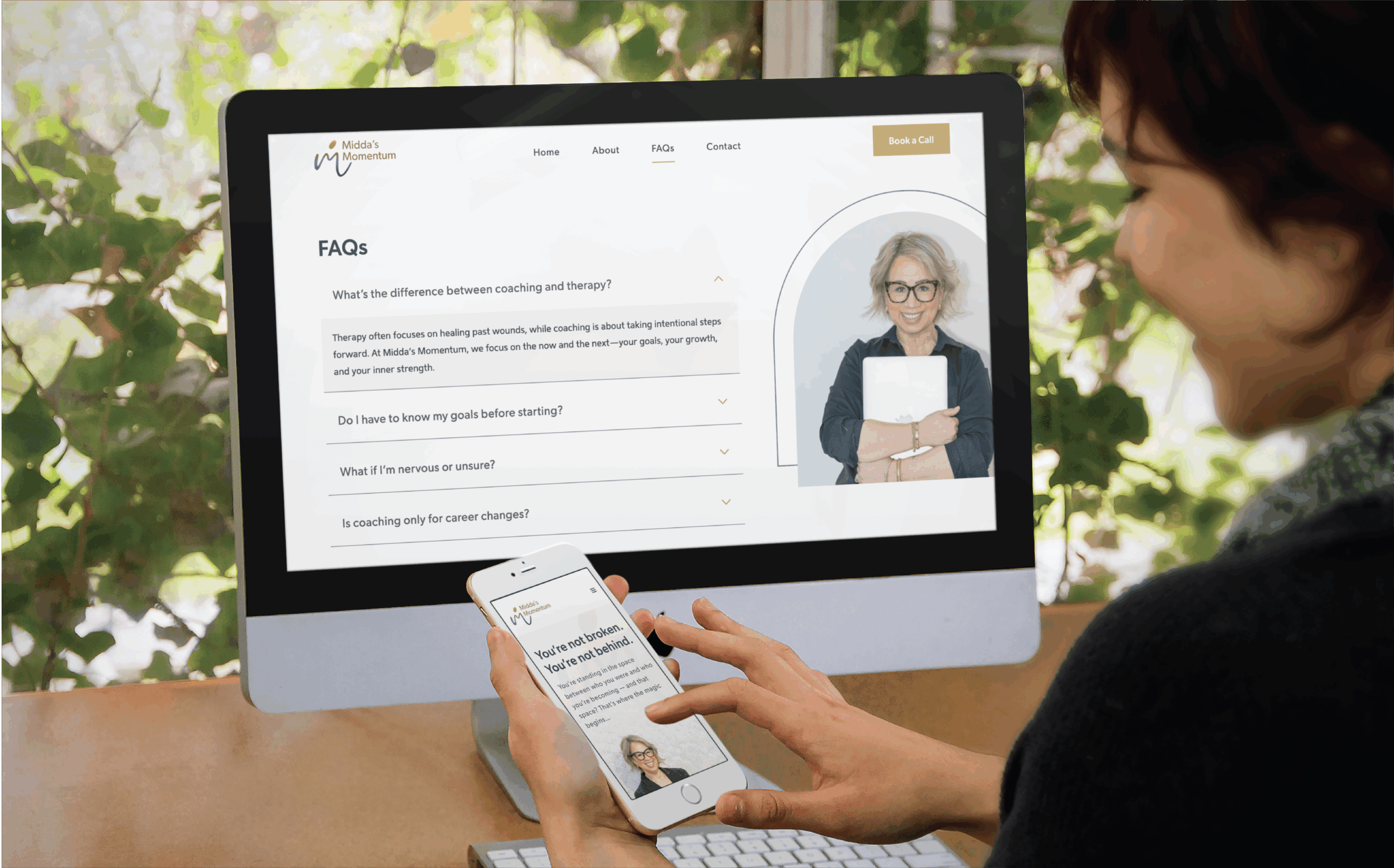 A person uses a smartphone and desktop computer to view an educational consultants’ website FAQ page, featuring a smiling woman with glasses holding a notebook, set against a leafy window background that reflects thoughtful website design.