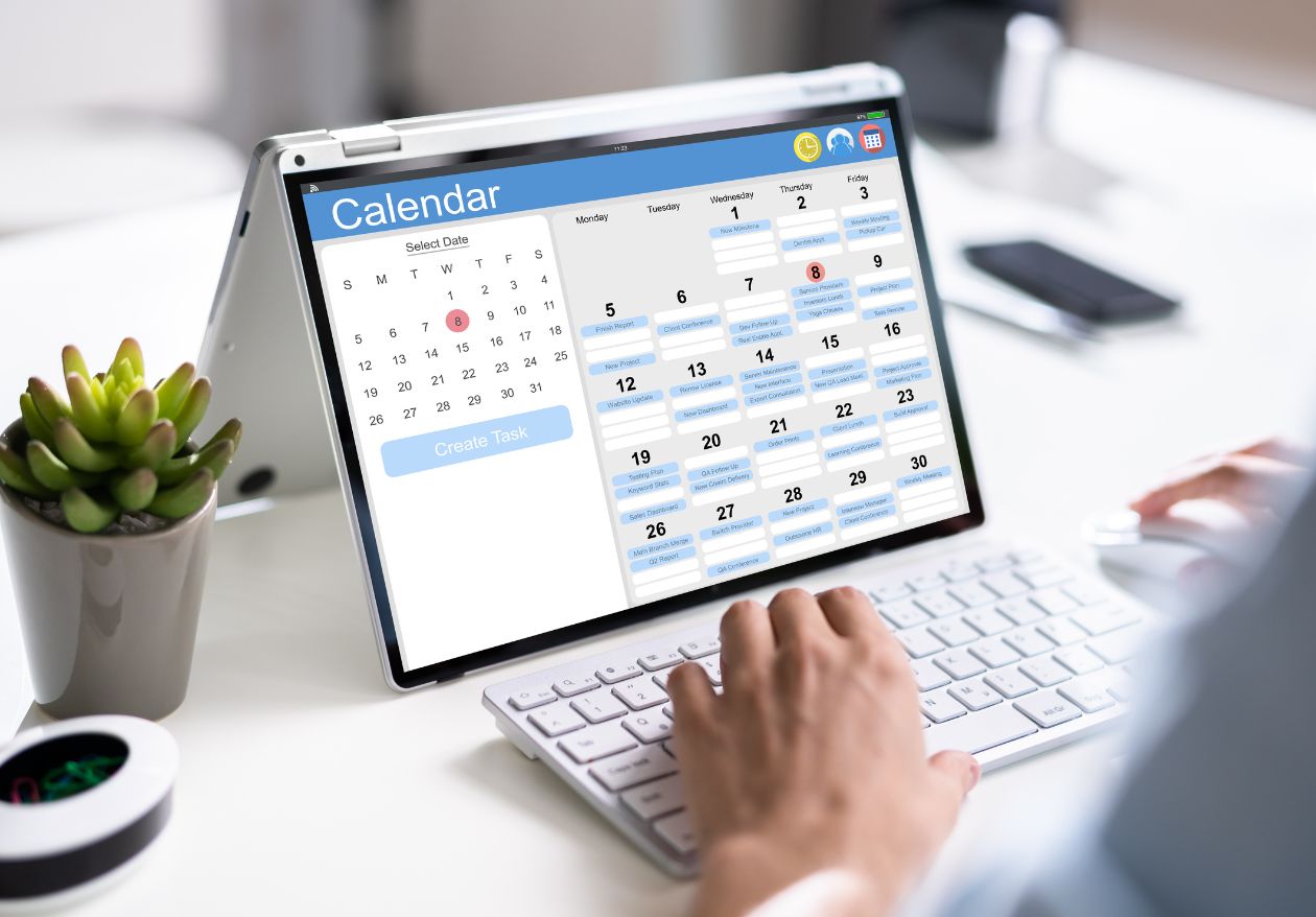 A person uses a computer displaying a digital calendar with task reminders. The workspace, perfect for moonlit media or web design services, includes a keyboard, mouse, and a small potted plant on a white desk.