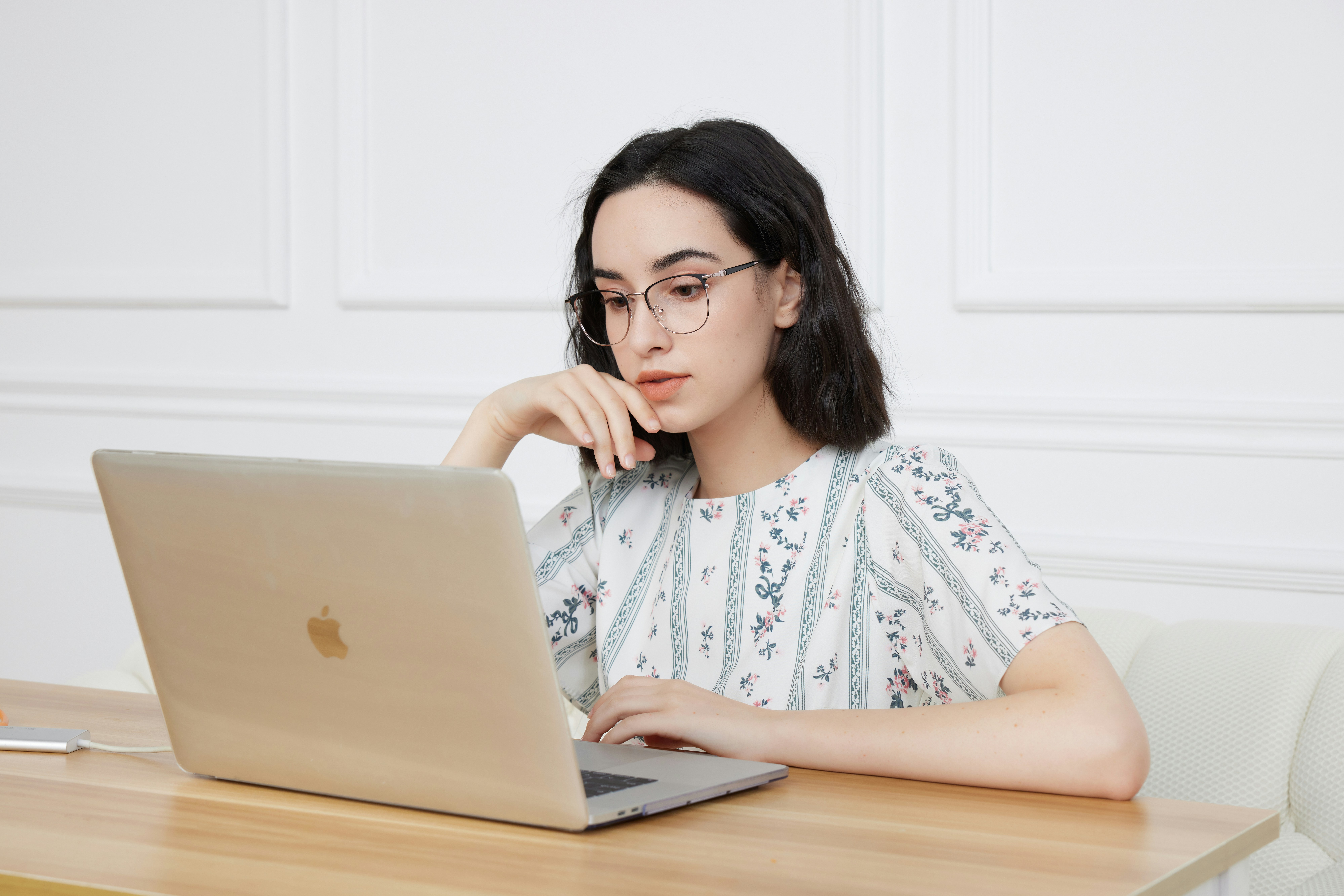 A young woman with dark hair and glasses sits at a wooden table, looking thoughtfully at an open laptop. She rests her chin on her hand, perhaps contemplating moonlit media or custom logo design. The background is a white wall.