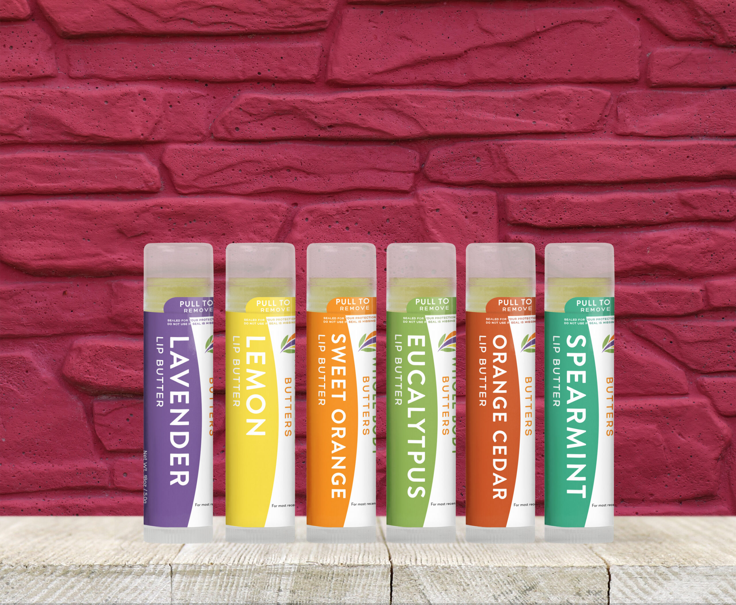 Six tubes of lip butter labeled Lavender, Lemon, Sweet Orange, Eucalyptus, Orange Cedar, and Spearmint are lined up on a white wooden surface in front of a brick wall painted dark pink—perfect inspiration for any web designer.