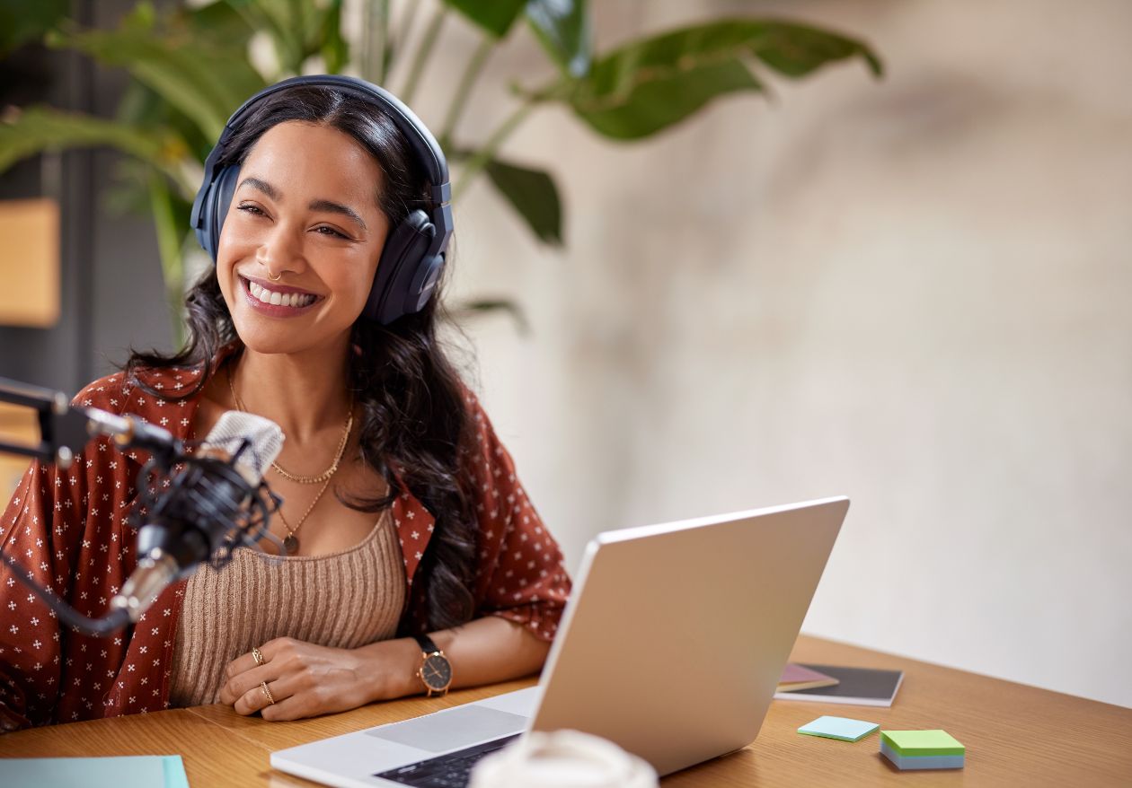 A smiling woman wearing headphones sits at a desk with a laptop, microphone, and colorful sticky notes, appearing to record a podcast or participate in an online meeting for Moonlit Media’s web designer and logo design services. Green plants are in the background.