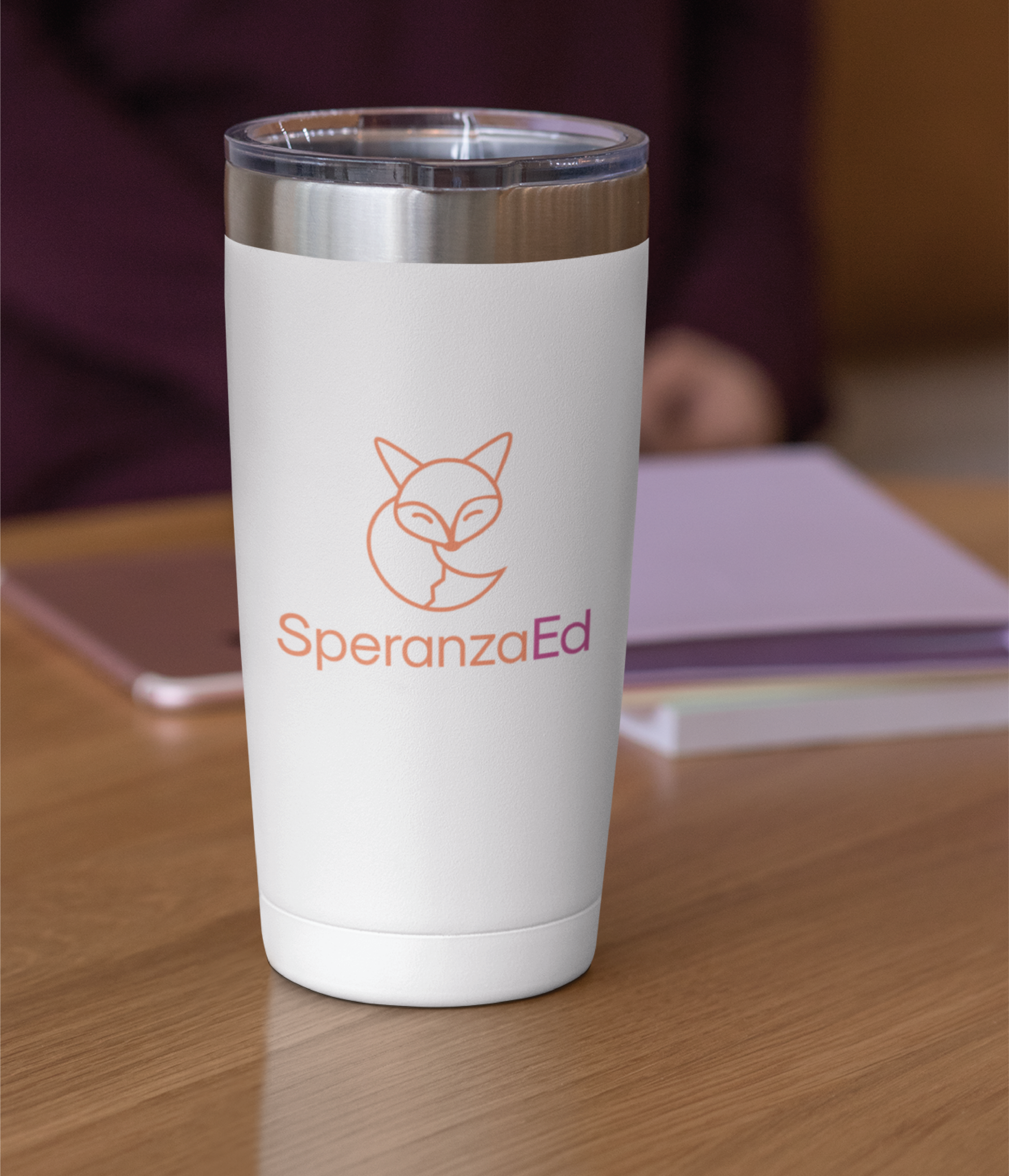 A white travel mug with a fox logo and the text SperanzaEd in orange and pink sits on a wooden desk, reflecting the branding style favored by educational consultants, next to a closed notebook and a blurred background.