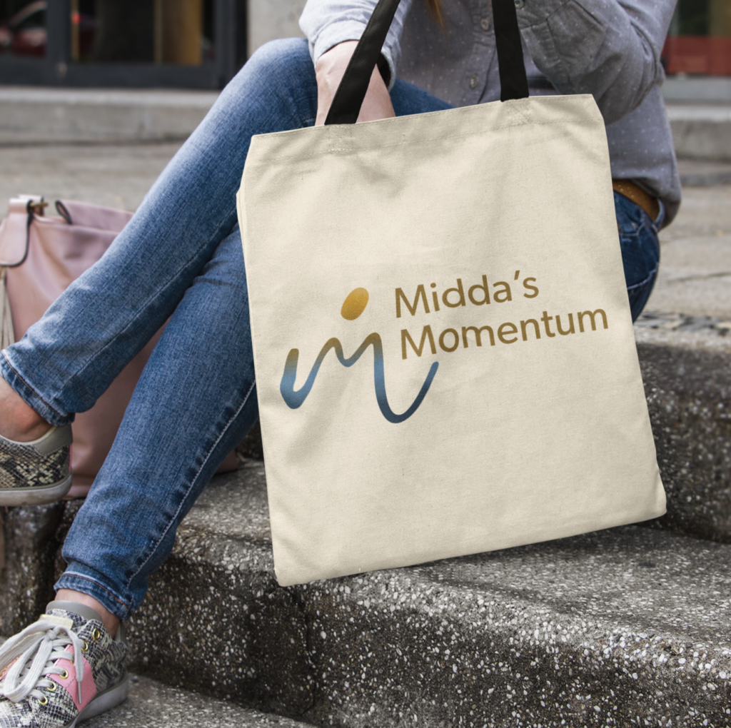 A person sits on outdoor steps holding a beige tote bag with Middas Momentum branding and a stylized “m” logo. The person wears jeans and colorful sneakers, while a light pink bag rests beside them.