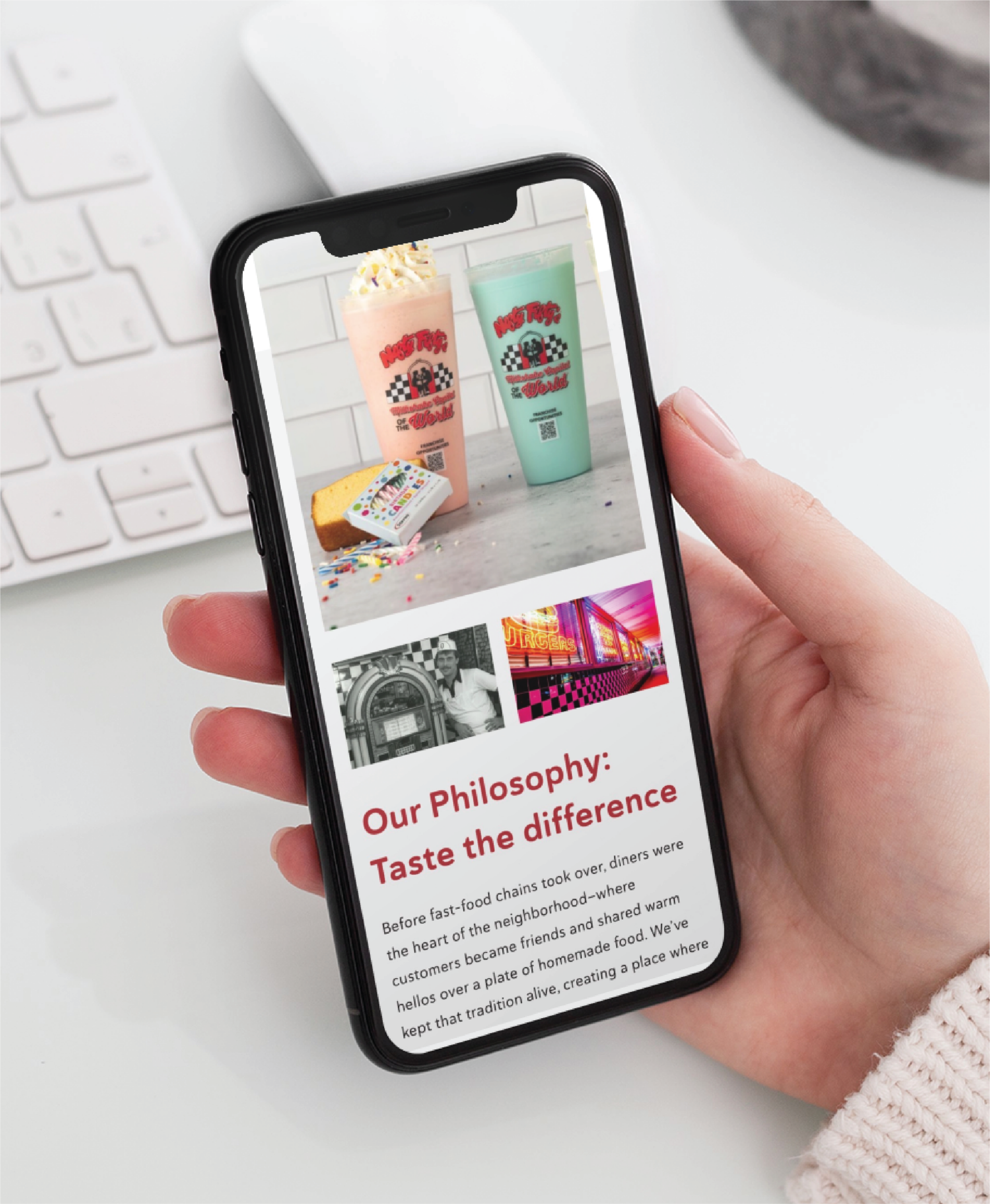 A hand holds a smartphone displaying a Philadelphia restaurant’s website design, featuring images of two colorful milkshakes, rainbow sprinkles, and cakes above text titled Our Philosophy: Taste the difference. A white keyboard is visible in the background.