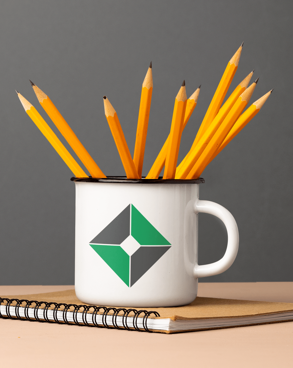 A white mug featuring a green and gray geometric logo design holds several sharpened yellow pencils. The mug sits on a closed spiral notebook with a brown cover against a plain gray background.