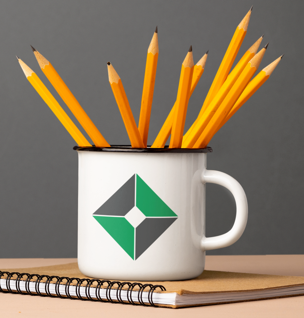 A white mug with a green and black geometric logo—perfect for showcasing your Logo Design skills—holds sharpened yellow pencils. The mug rests on a spiral notebook with a brown cover, set against a plain gray background.