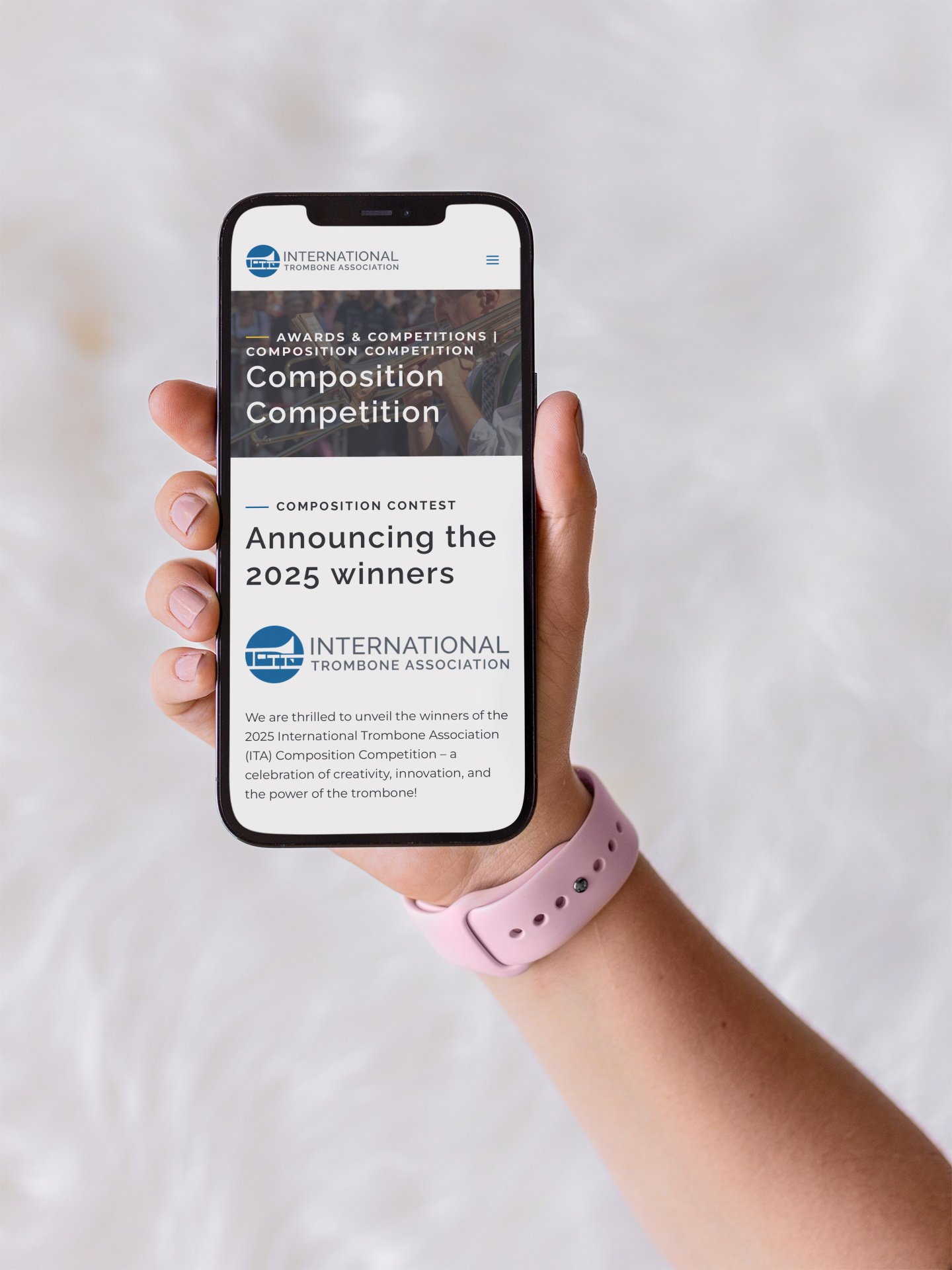 A hand wearing a light pink smartwatch holds a smartphone displaying a moonlit media webpage announcing the 2025 winners of the International Trombone Association’s Composition Competition.