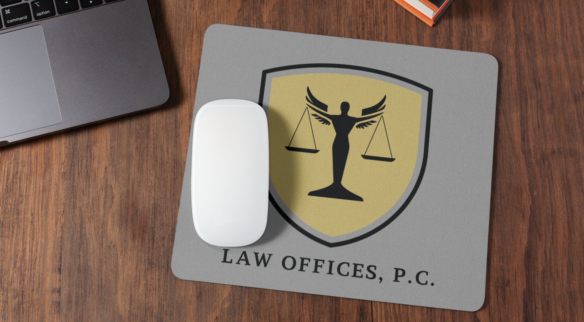 A white mouse sits on a gray mouse pad with a gold and black law office logo design featuring Lady Justice. The text below reads “LAW OFFICES, P.C.” The pad rests on a wooden desk beside a laptop.