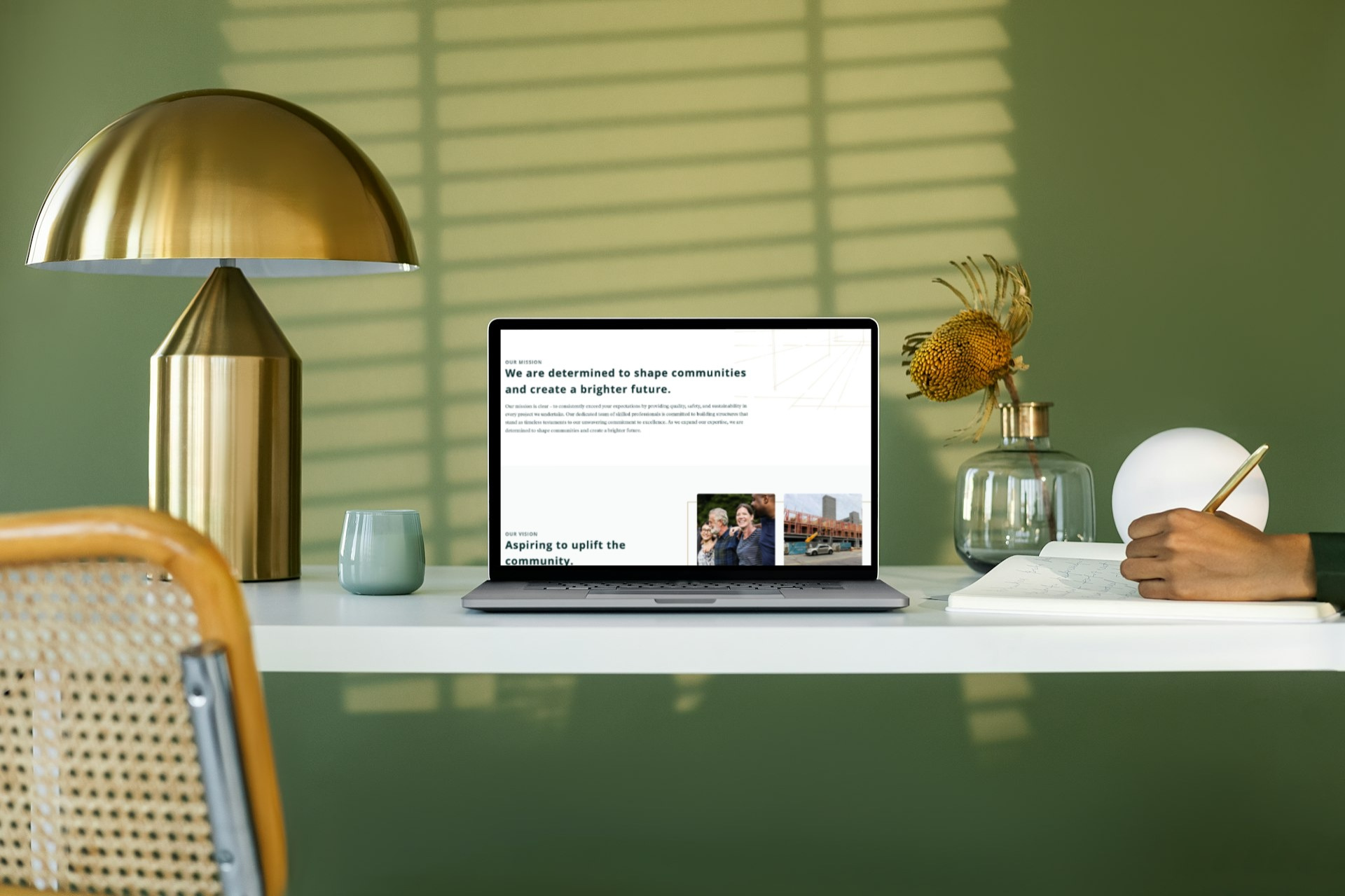 A modern desk setup by Moonlit Media features a gold lamp, open laptop displaying web design services, a glass vase with dried flowers, and a person writing in a notebook—all set against a green wall with shadowed blinds.