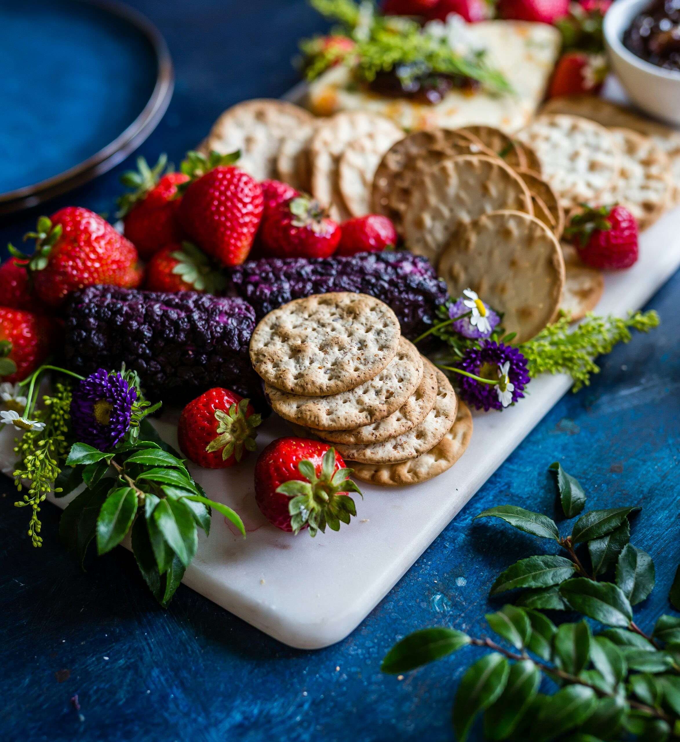 A marble platter with assorted crackers, fresh strawberries, and a log of berry-covered cheese, garnished with flowers and greenery, styled as artfully as our custom website design creations—displayed on a blue surface.