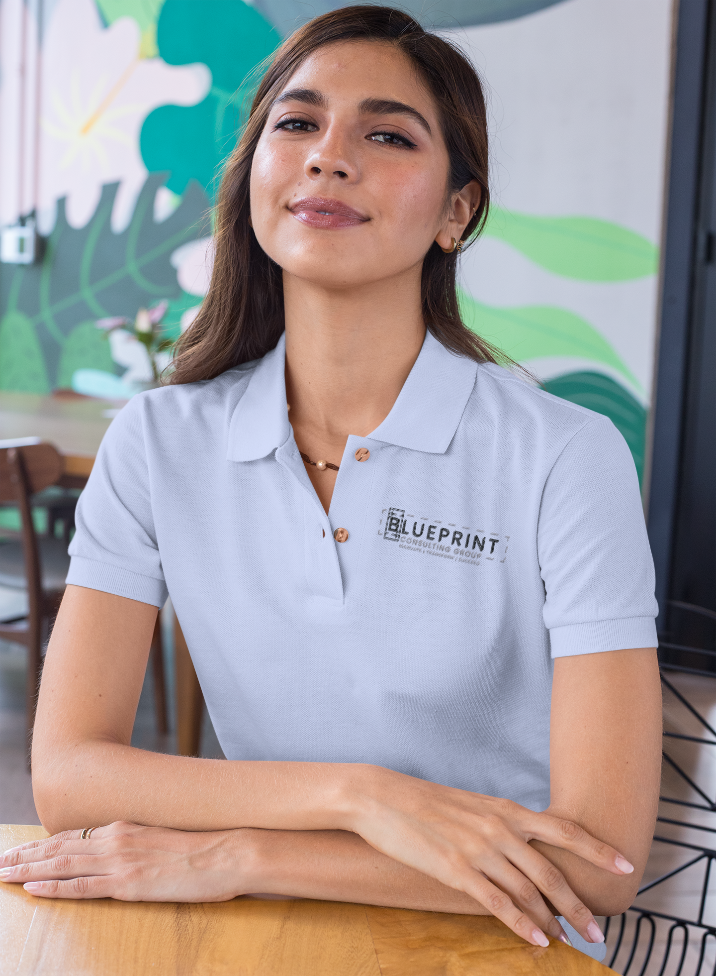 A woman with long brown hair sits at a table in a modern, brightly decorated café, wearing a light blue polo shirt with the BLUEPRINT logo on it. She smiles gently and looks confidently at the camera, representing a leading consulting group.