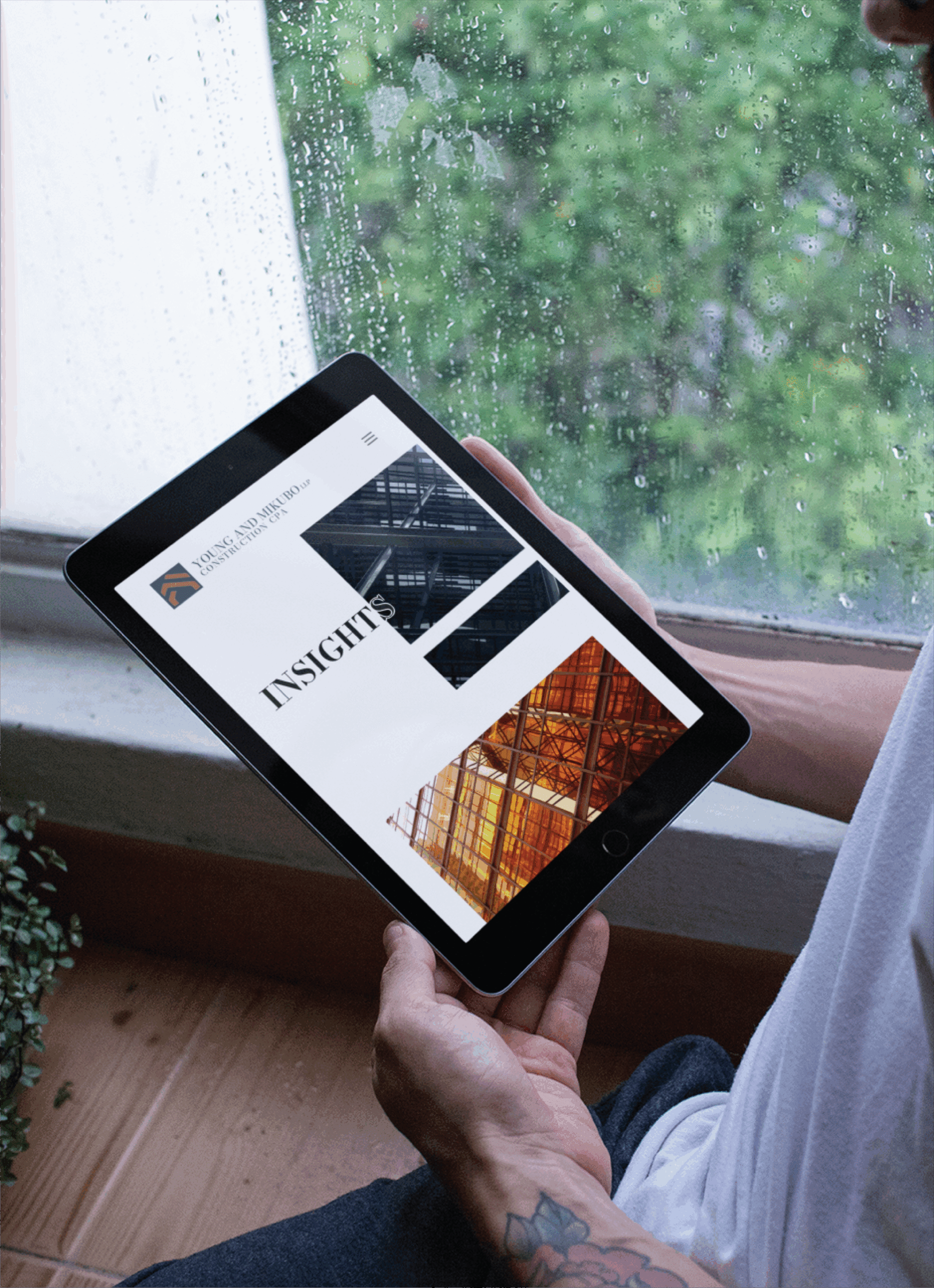 A person holding a tablet displaying a roofing company’s website design with architectural images and the word INSIGHTS, sitting by a rainy window.