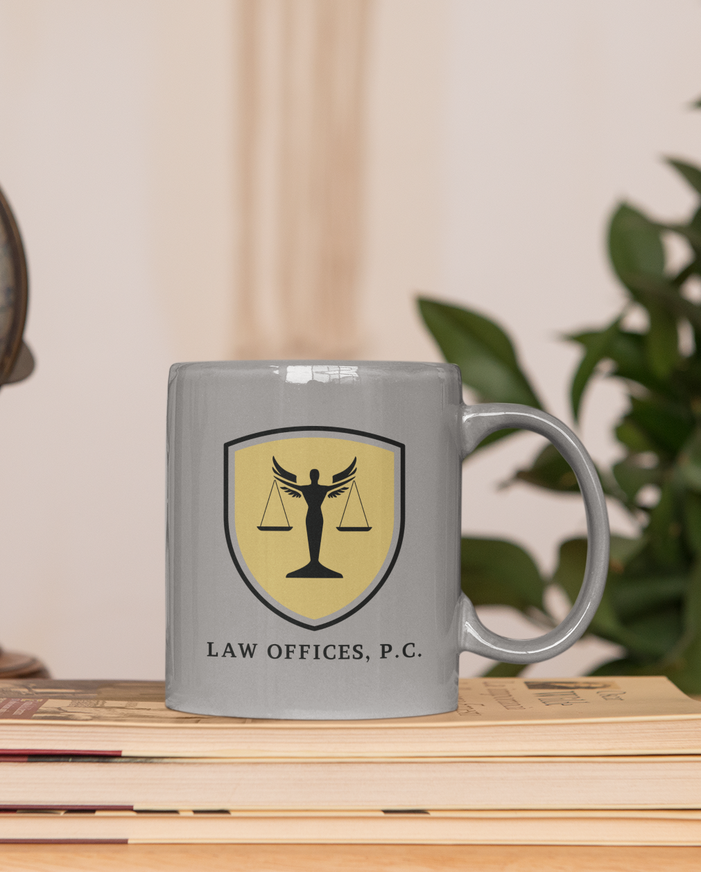 A gray mug with a yellow and black law office logo, ideal for showcasing your law offices Atlanta branding or LOGO design. "LAW OFFICES, P.C." is printed below the logo. The mug rests on stacked books beside a green plant.