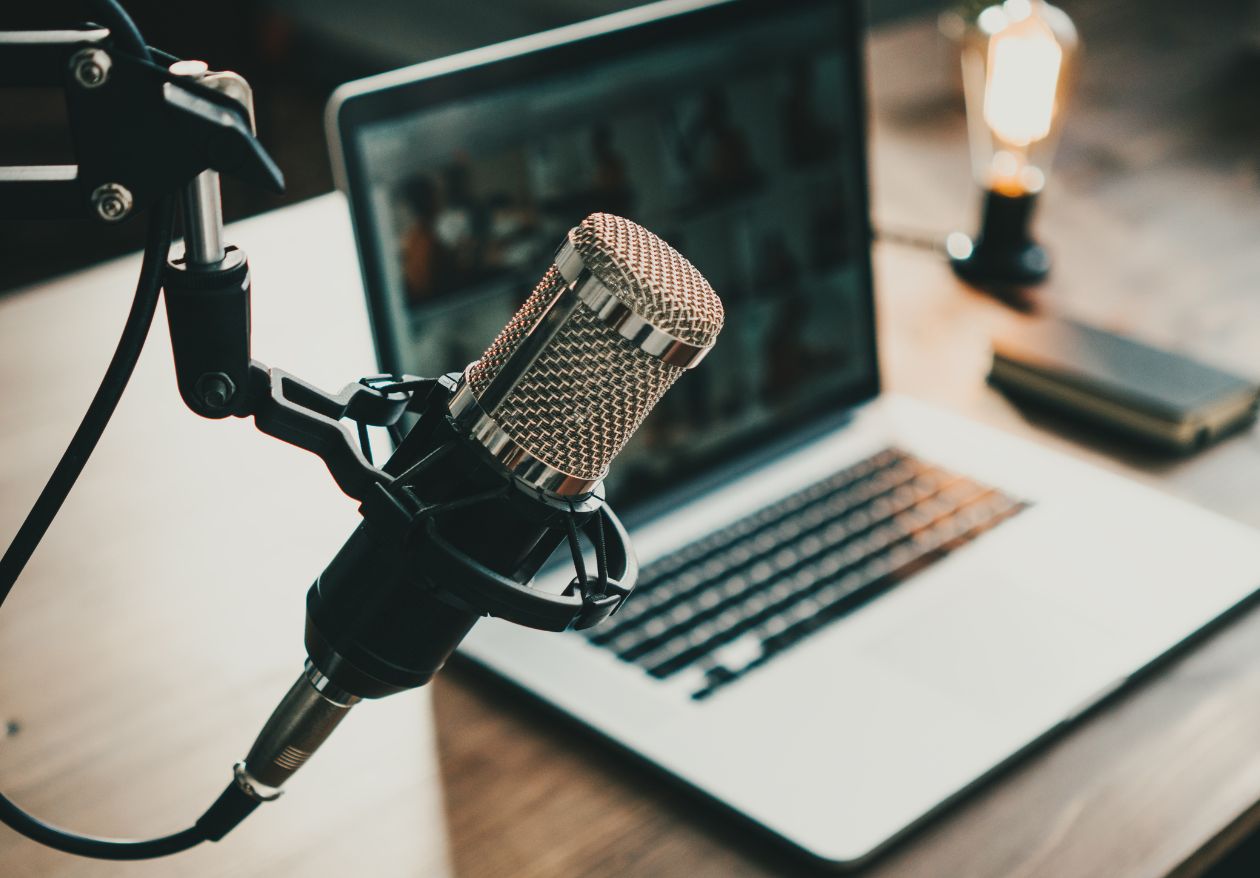 A close-up of a studio microphone on a stand in front of an open laptop, with a blurred background including a notebook, a small lamp, and branded materials showcasing custom logo design by Moonlit Media on a wooden desk.