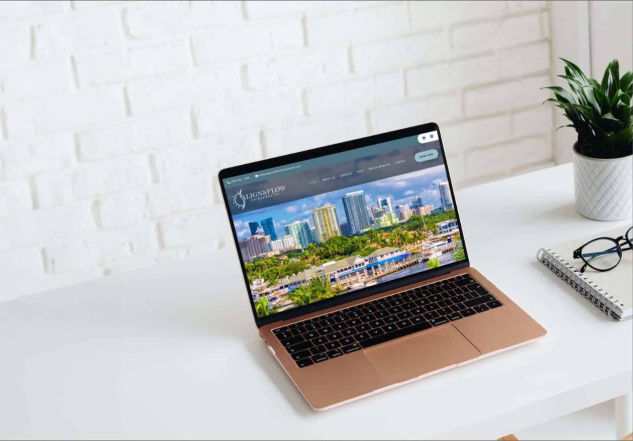 A gold laptop displaying a cityscape website sits on a white desk next to a small potted plant and spiral notebook, perfect for showcasing custom logo design by Moonlit Media, with a white brick wall in the background.