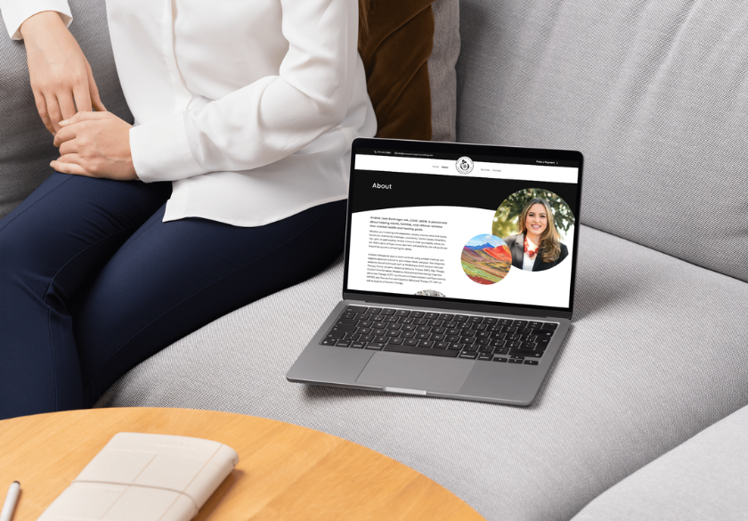 A person sits on a light gray couch next to an open laptop displaying website bio content with a photo of a woman and text. A closed notebook and pen rest on the round wooden table in front.