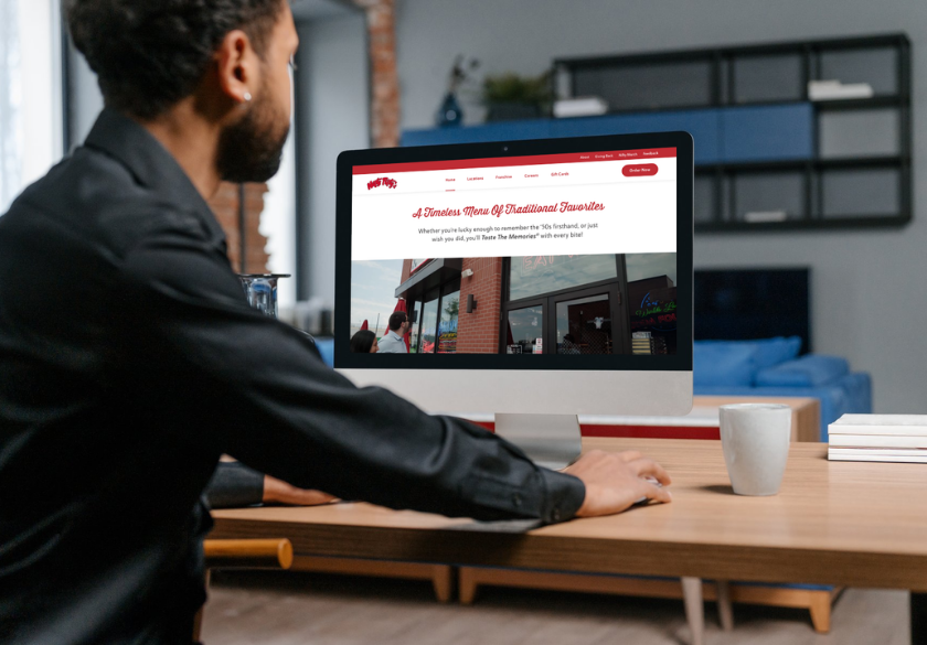 A person sits at a desk looking at a small business website on their computer, which shows a red and white header and an image of a storefront. A coffee mug and notebook rest beside them in a modern living room, suggesting they’re planning the number of website pages.