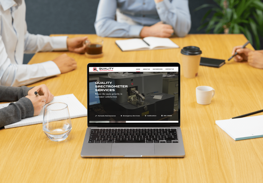 A laptop displaying a Quality Spectrometer Services small business website is on a wooden table, surrounded by glassware, notebooks, pens, a coffee cup, and people sitting and taking notes.