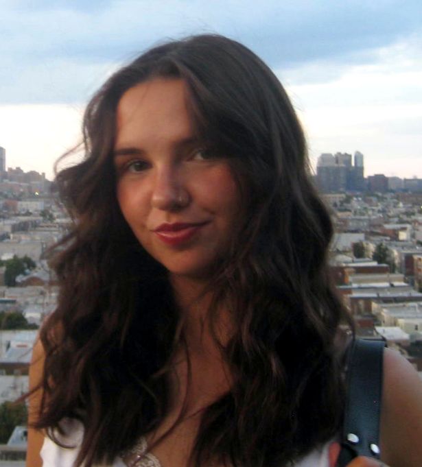 A young woman with long wavy brown hair smiles softly at the camera. She is outdoors, with a cityscape backdrop—an inspiring face behind this woman-owned branding studio, ready to transform ideas with creativity and passion.