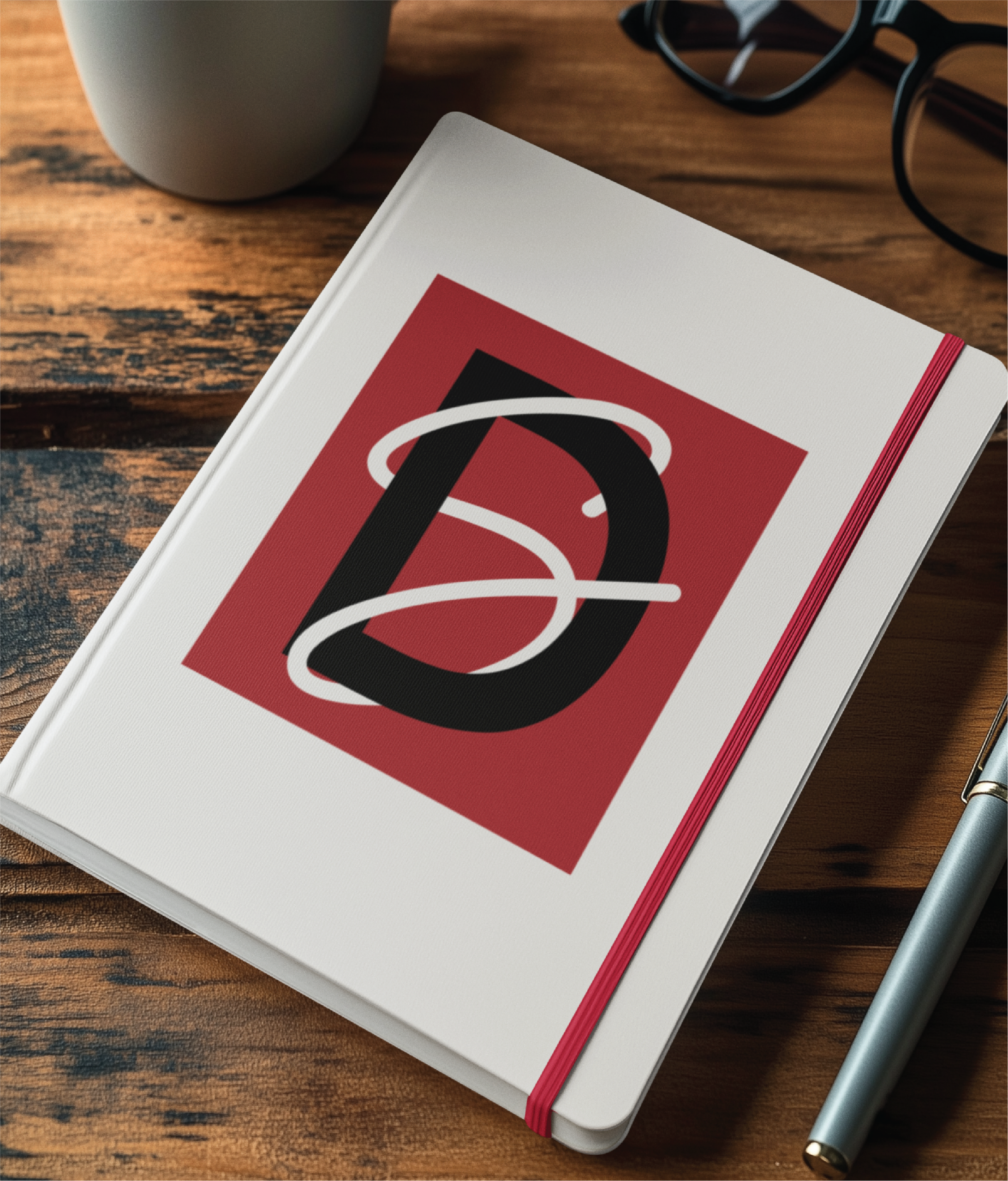A white notebook with a red elastic band, featuring a bold “D” logo on the cover, sits on a wooden desk near a pen, black glasses, and a white mug—perfect for sketching custom website design ideas in Philadelphia PA.