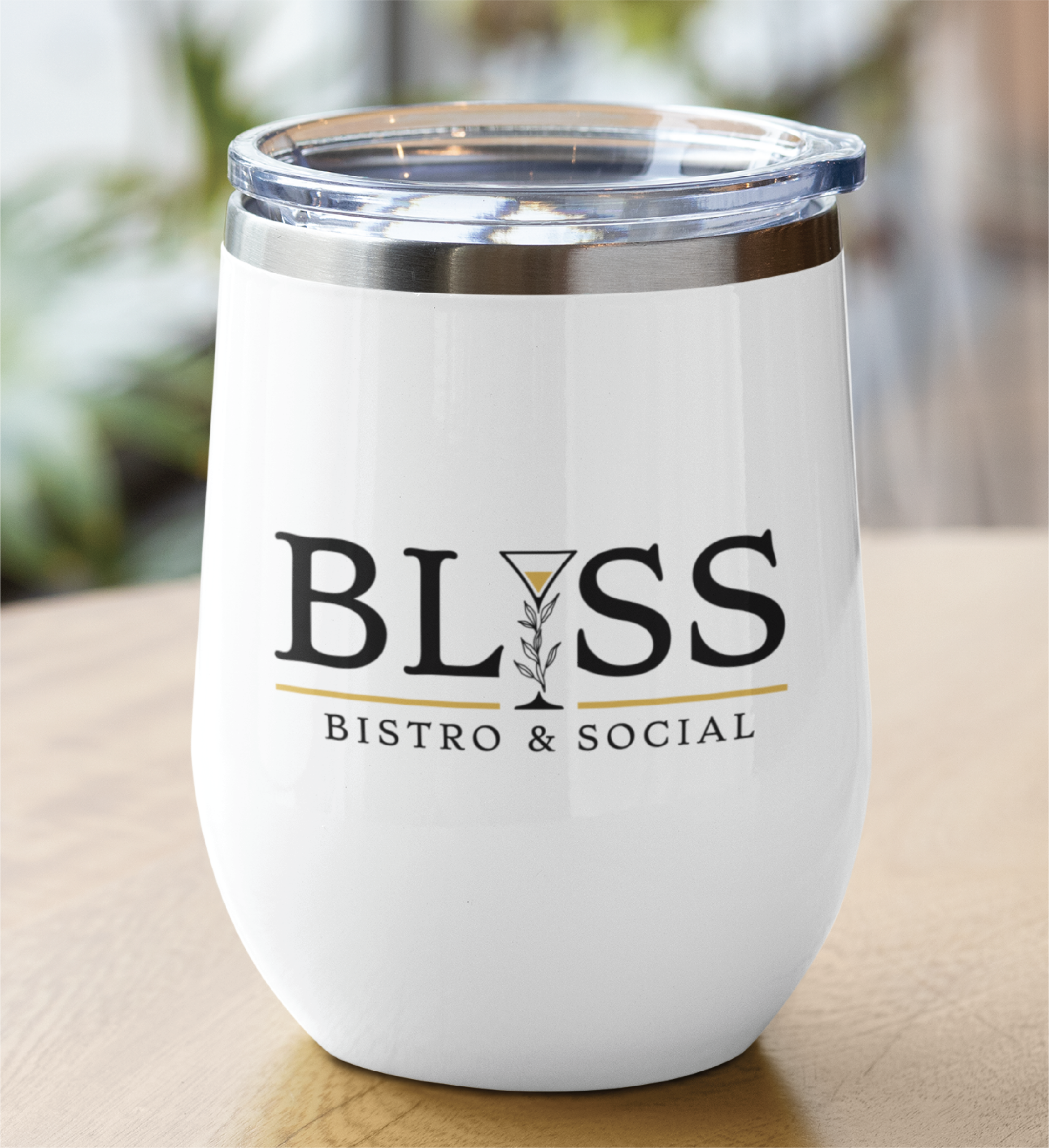 A white insulated tumbler with a clear lid sits on a wooden table. The tumbler displays the restaurant's custom logo design, “BLISS BISTRO & SOCIAL,” with a martini glass as the “I” in “BLISS”—a chic touch for any Florida eatery.