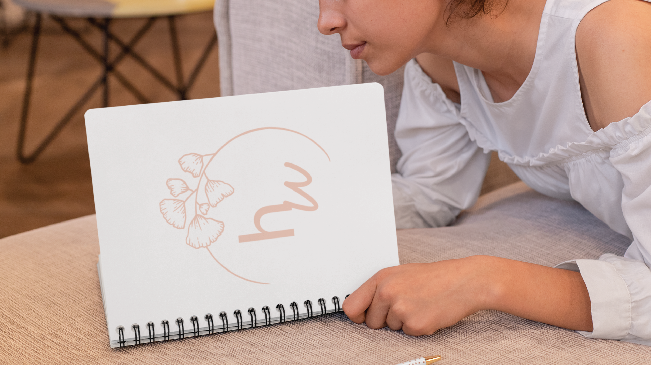 A person in a white shirt holds and looks at an open spiral notebook displaying a minimalistic Custom Logo Design of leaves and the cursive letters hw inside a circular line.