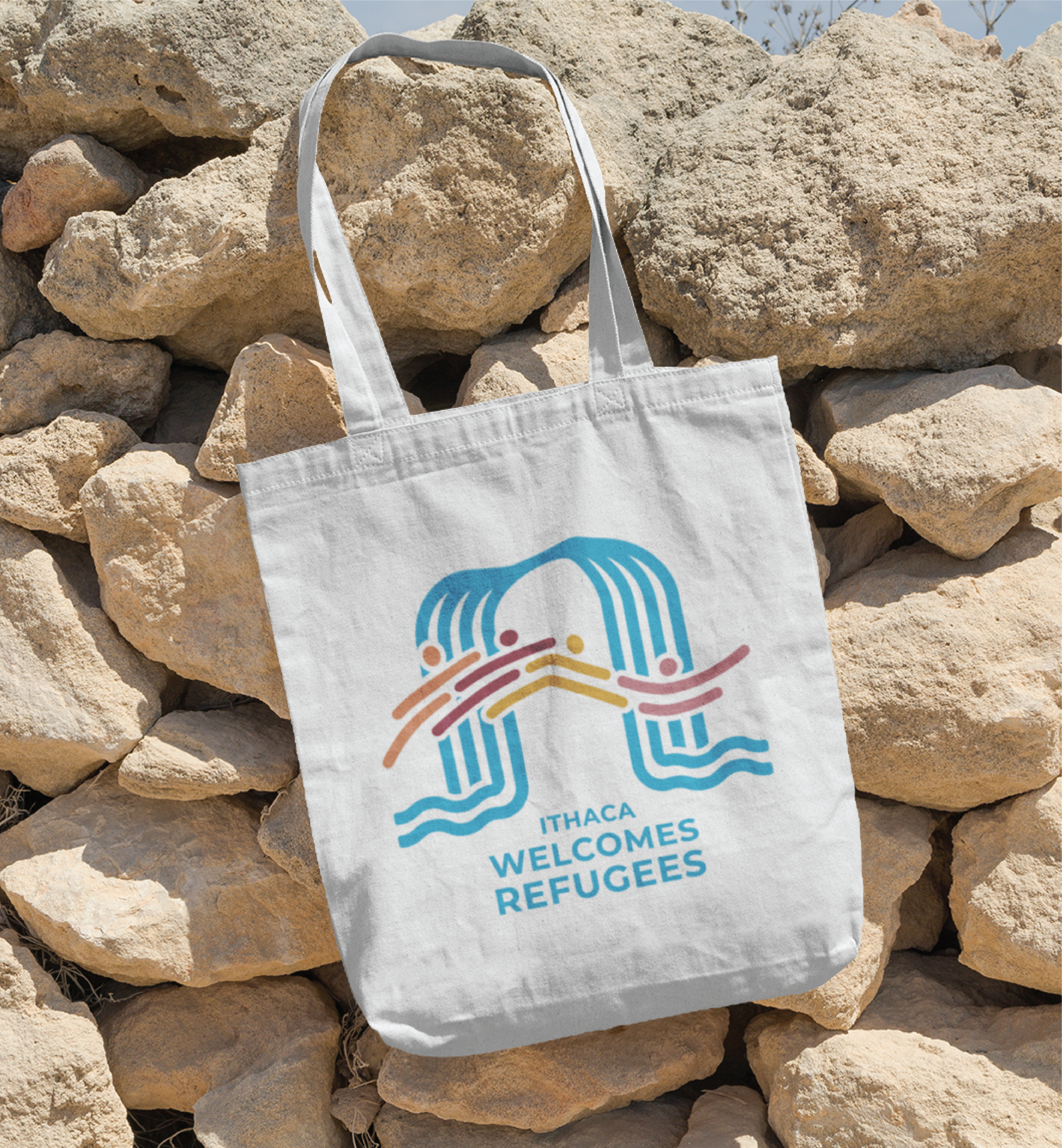 A white tote bag hangs on a stone wall, displaying a colorful print design with stylized figures and water, and the text Ithaca Welcomes Refugees in blue—a cheerful accessory that would fit right in at a Pennsylvania market.