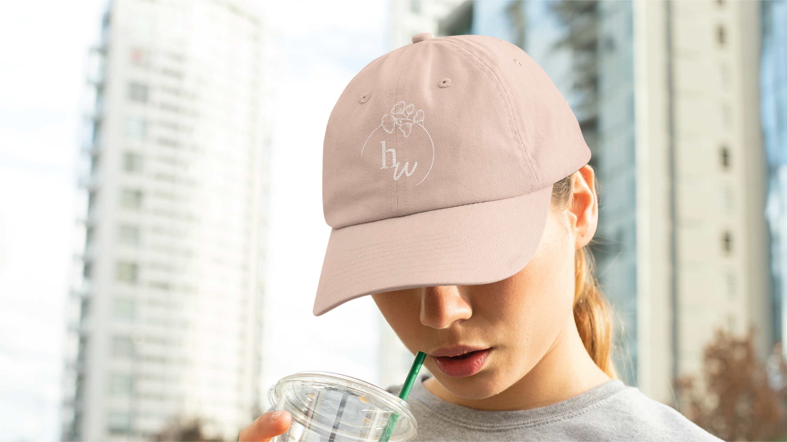A person wearing a light pink baseball cap with an embroidered flower and custom logo design sips a drink through a green straw, with New York’s tall buildings blurred in the background.