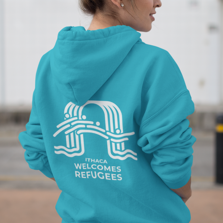 A person wearing a bright blue hoodie featuring a white print design and text that reads Ithaca Welcomes Refugees on the back. Outdoors in Pennsylvania, they’re turned slightly away from the camera.