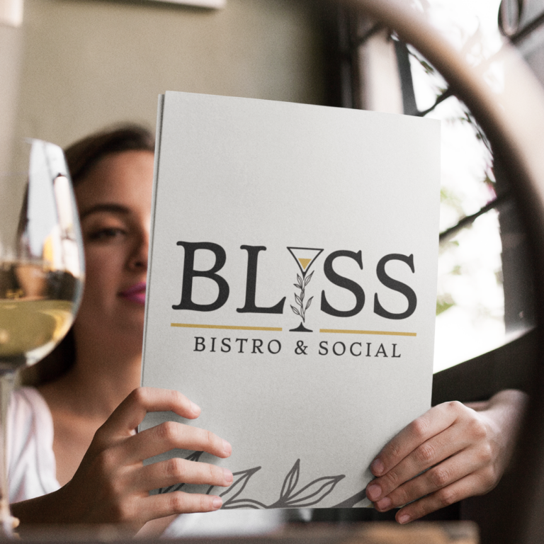 A woman holds a menu featuring a custom logo design with “BLISS Bistro & Social” on the cover, sitting at a table with a glass of white wine in a cozy, sunlit Florida restaurant.