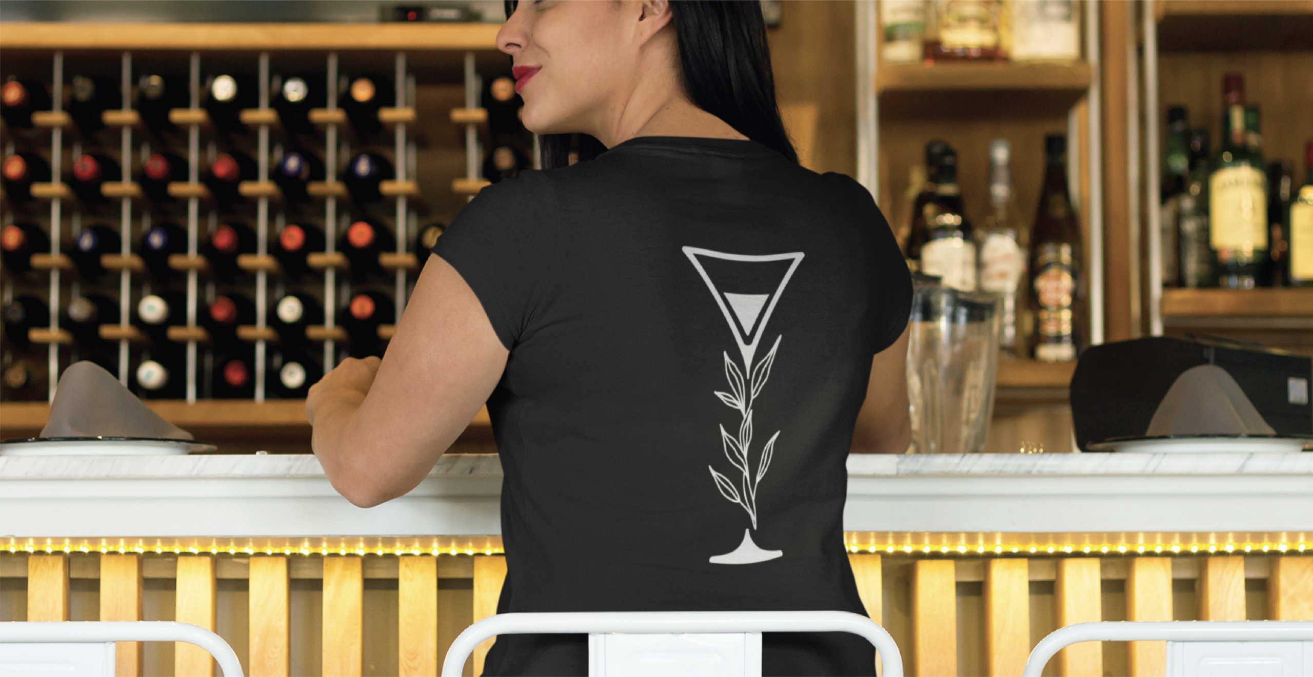 A woman sits at a bar in a Florida restaurant, wearing a black shirt featuring a custom logo design: a white outline of a cocktail glass with a leafy accent, with a wine rack displayed in the background.