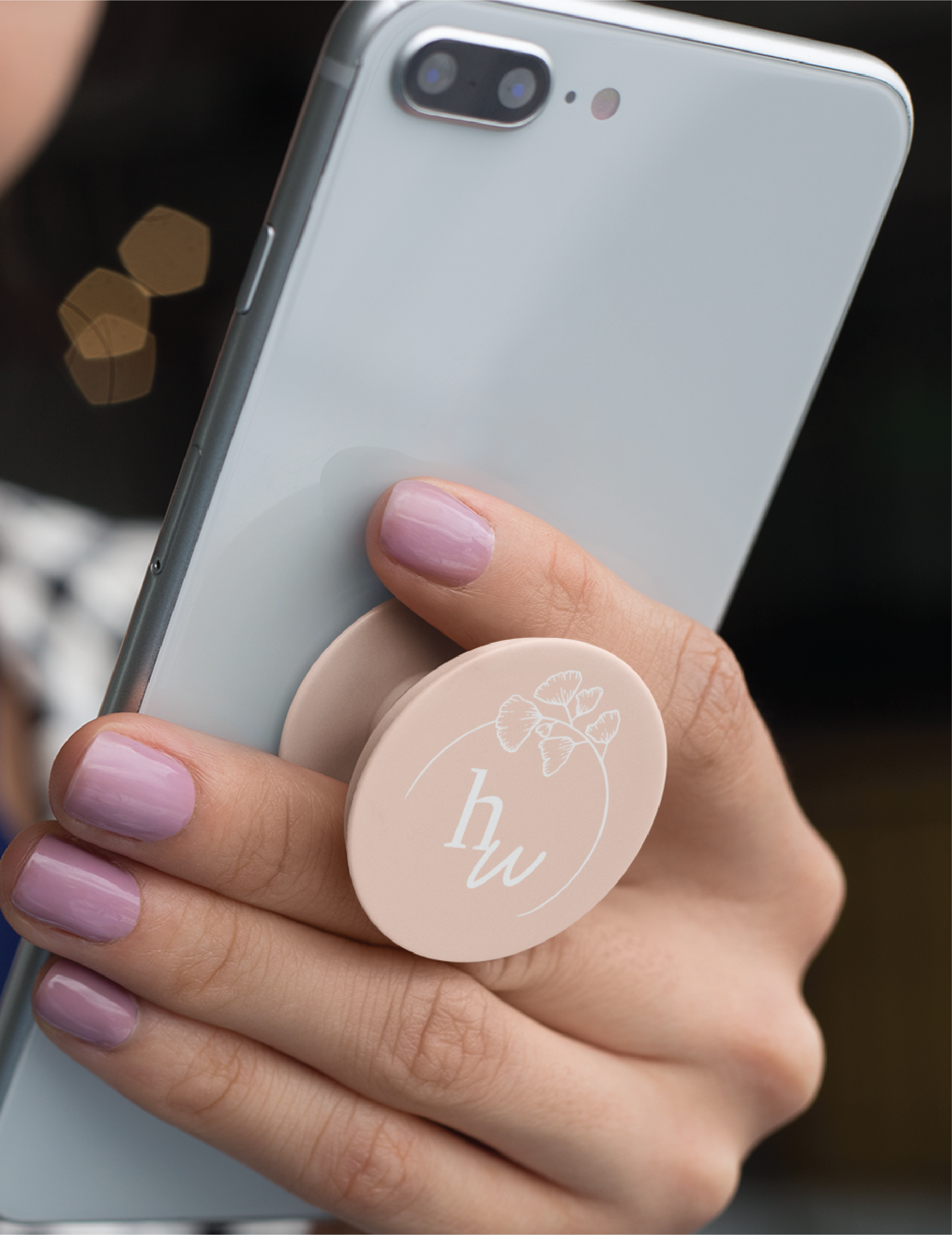 A hand with light purple nail polish holds a smartphone with a pale pink pop socket featuring a custom logo design—floral details and the letters hw in white, inspired by a New York nonprofit organization.