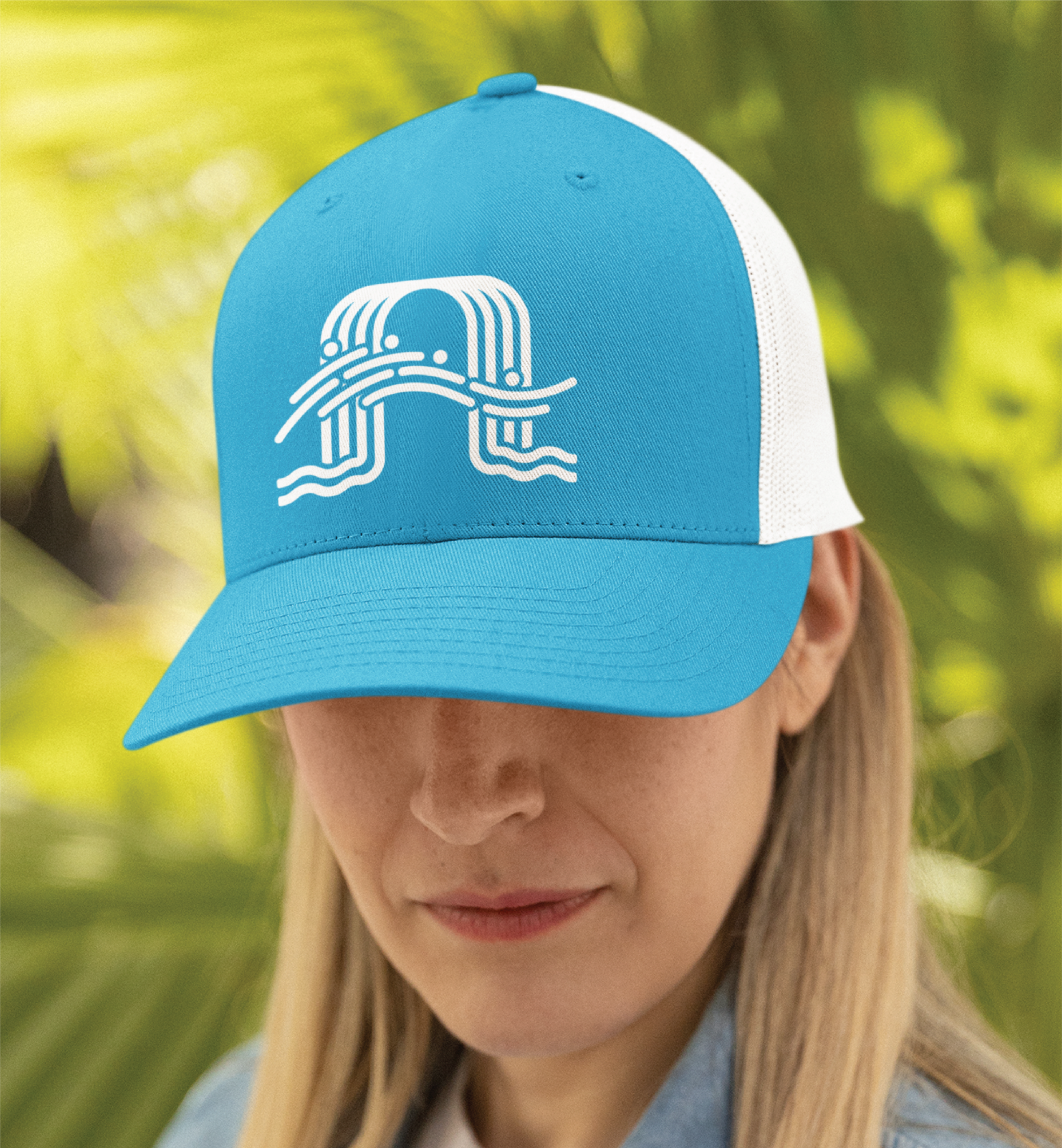A woman with long blonde hair wears a blue and white mesh baseball cap featuring a stylized print design on the front. The background is filled with green, leafy plants.