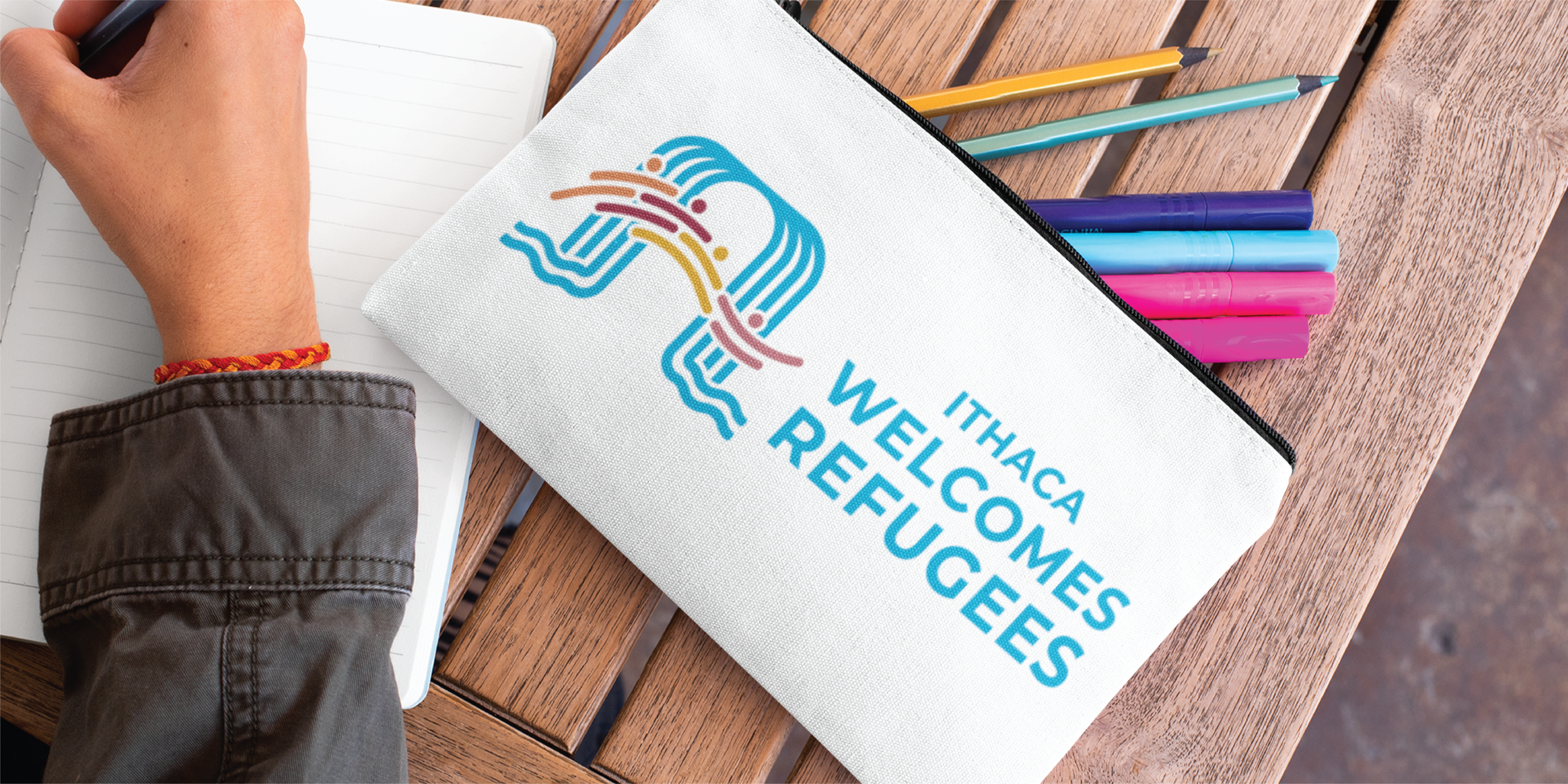 A person writes in a notebook next to a white pouch with colorful pens, featuring “Ithaca Welcomes Refugees” and a stylized graphic of two birds—a touch reminiscent of Pennsylvania’s vibrant print design traditions.