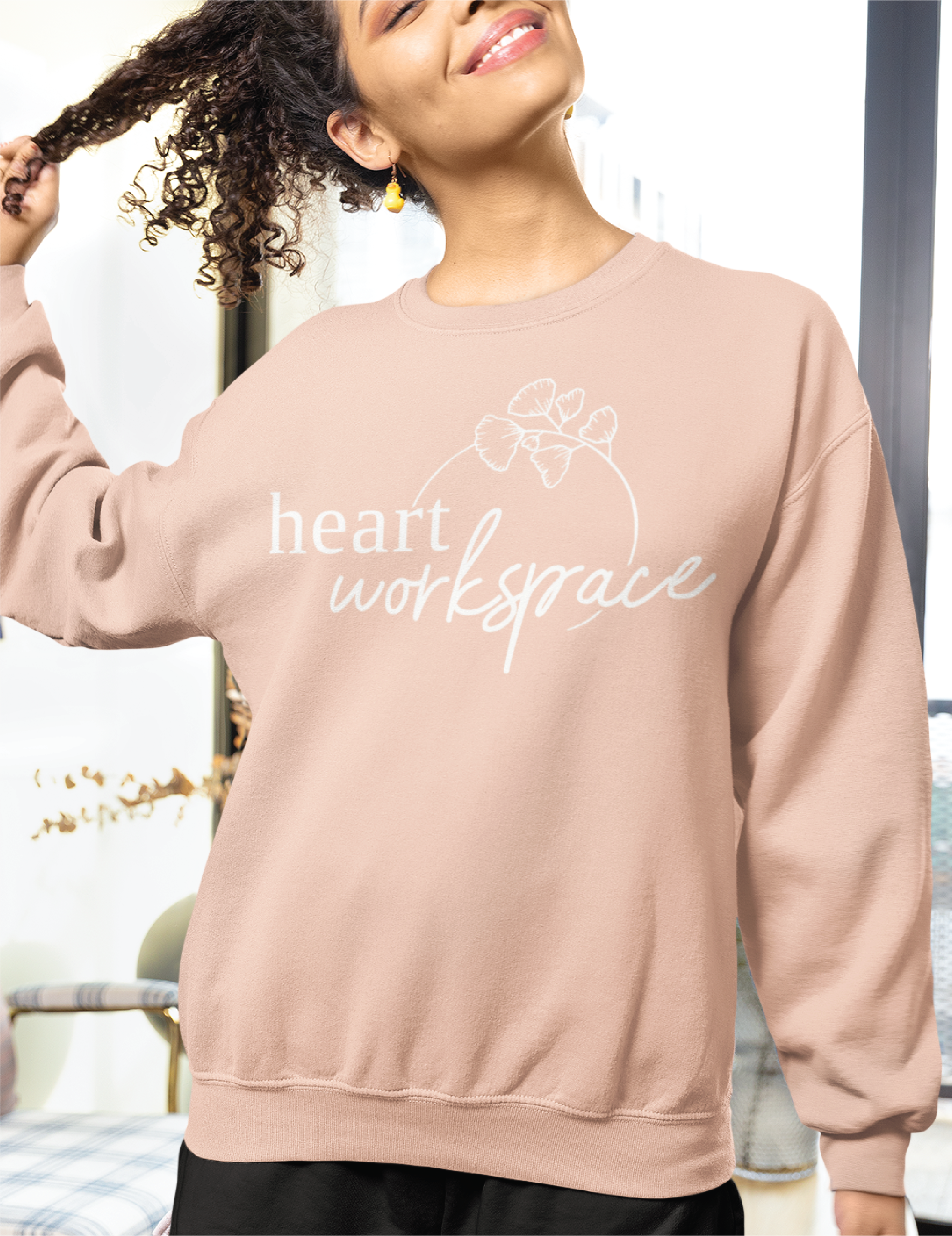 A woman smiles and lifts her curly hair while wearing a light pink sweatshirt that reads heart workspace with a small graphic of leaves—a custom logo design for a nonprofit organization. She stands in a bright, cozy room with modern decor.