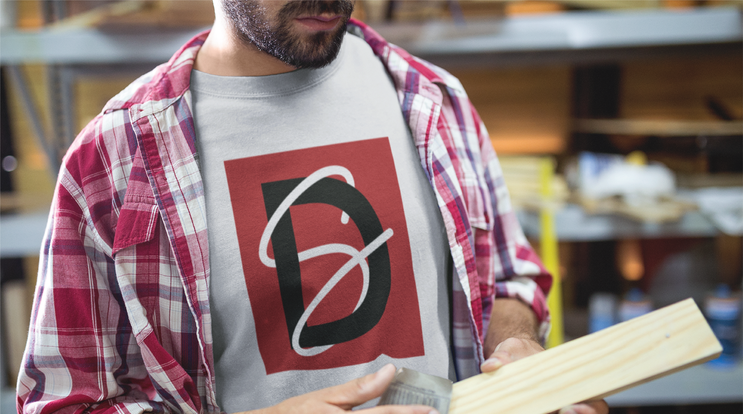 A person in a plaid shirt over a t-shirt with a bold red and black graphic design holds a piece of light-colored wood in a Philadelphia workshop, possibly crafting decor for a 1950s-themed diner or elements for custom website design projects.