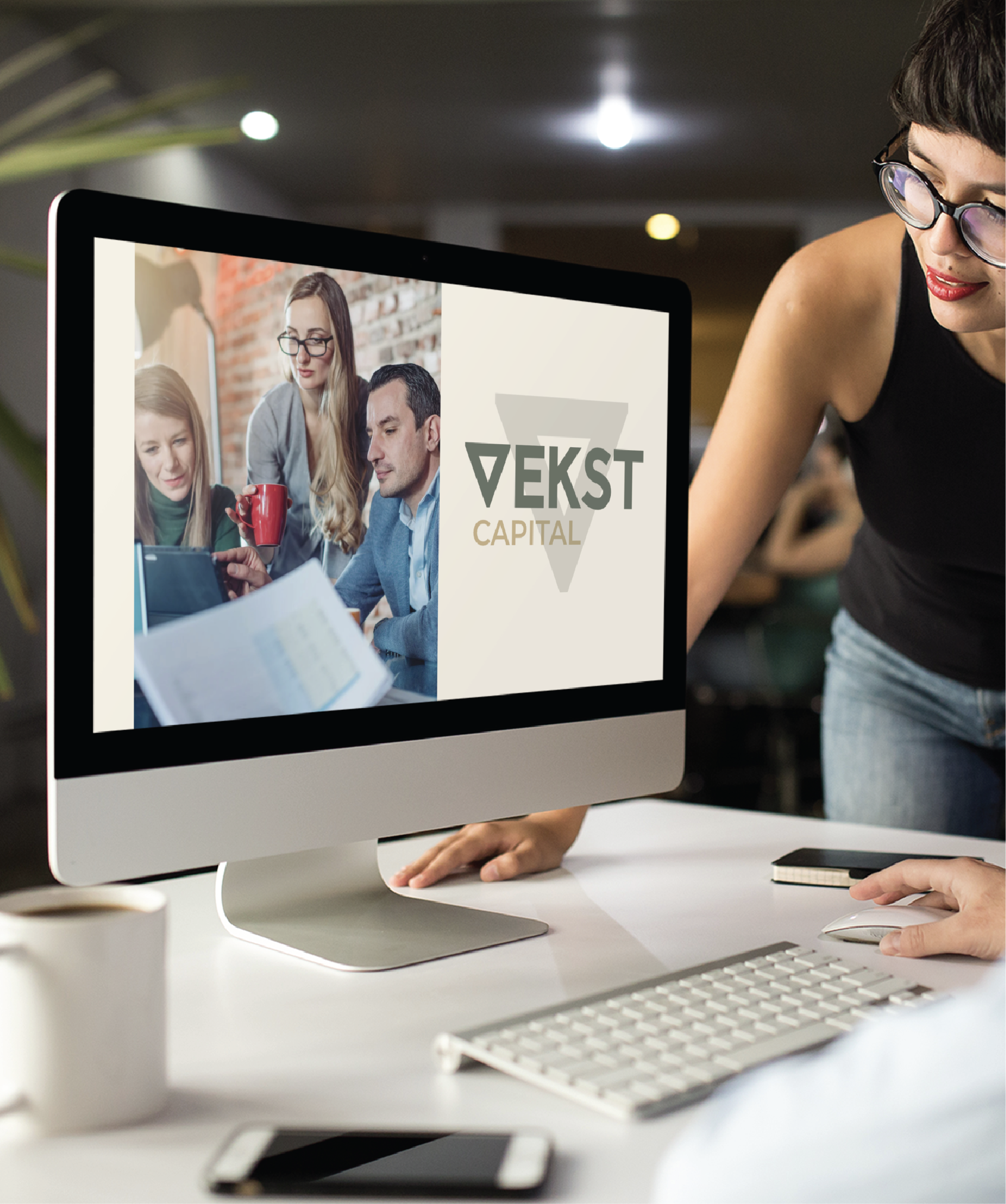 A person standing at a desk points to an iMac screen displaying a group collaborating under the text “VEKST CAPITAL.” A keyboard, coffee cup, and hand using a mouse add to the workspace—perfect inspiration for web design teams in South Carolina.