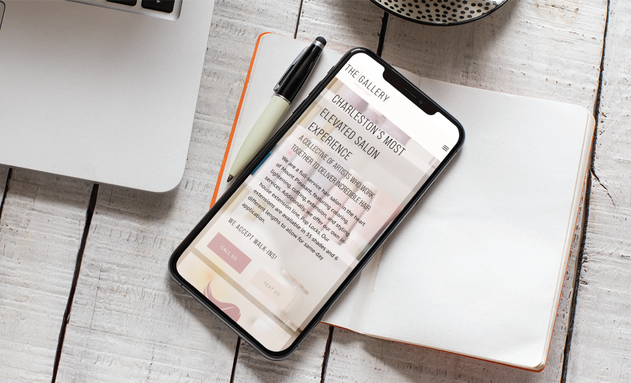 A smartphone displaying a Beauty Salon website rests on an open blank notebook with a pen, next to a laptop and a dotted bowl on a white wooden table in South Carolina.