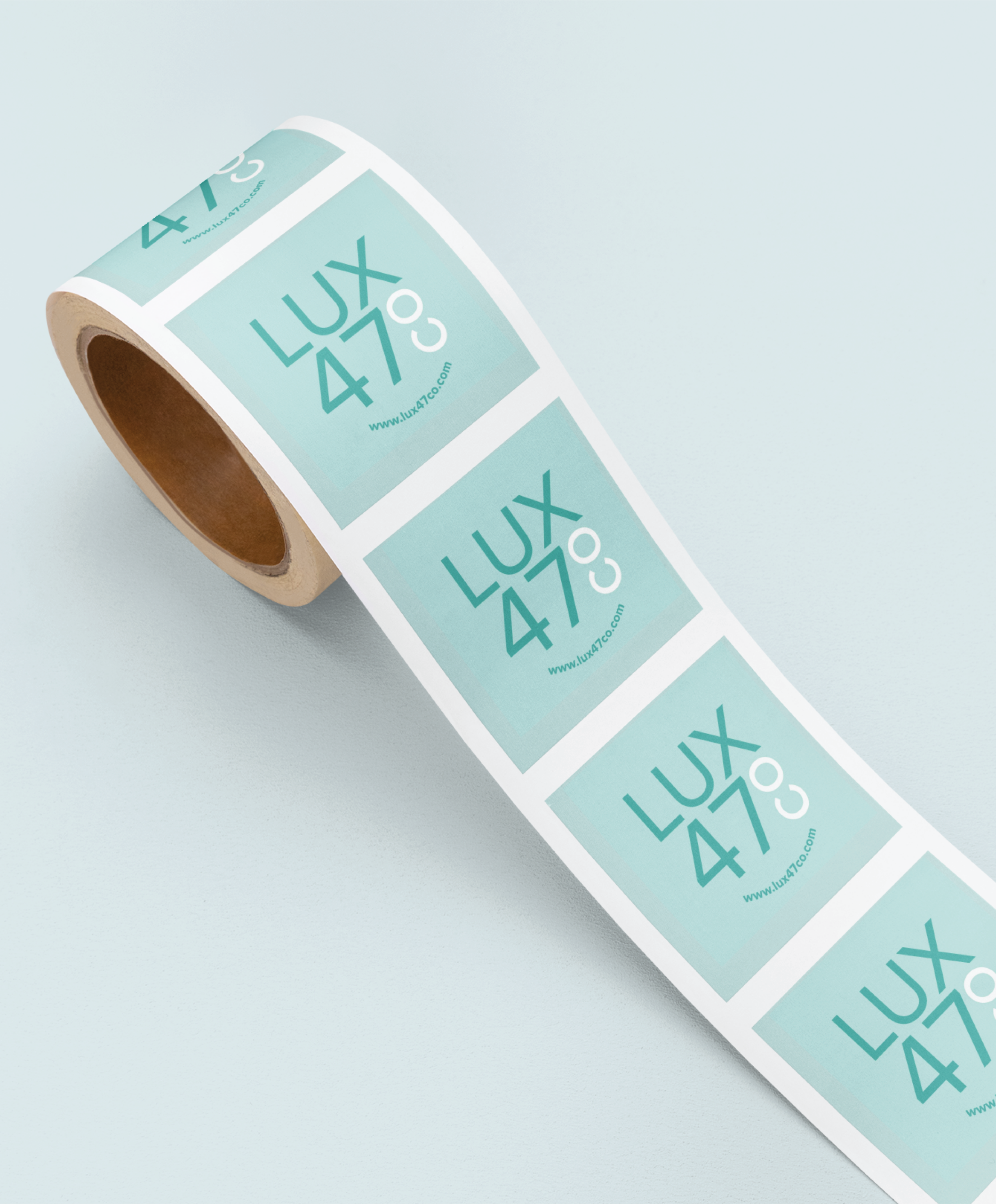 A roll of rectangular stickers with a teal background, displaying “LUX 47°C” and a website link, showcases premium print design—ideal for handcrafted soap makers in Pennsylvania—against a light blue surface.