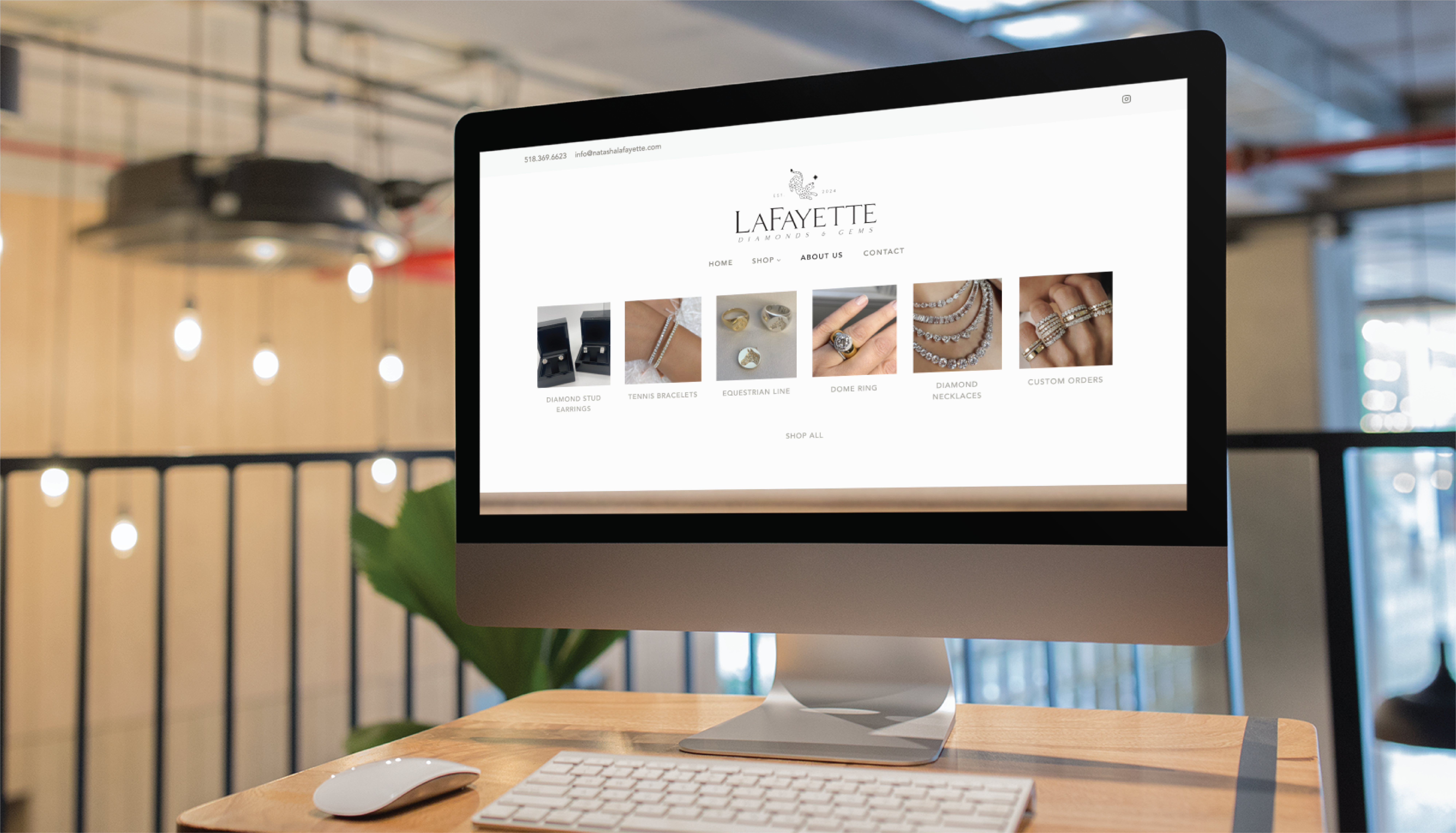 A desktop computer on a wooden desk displays the Lafayette Jewelry website, showcasing custom website design and jewelry images. The background features modern office decor with hanging lights and a plant in a chic New York setting.