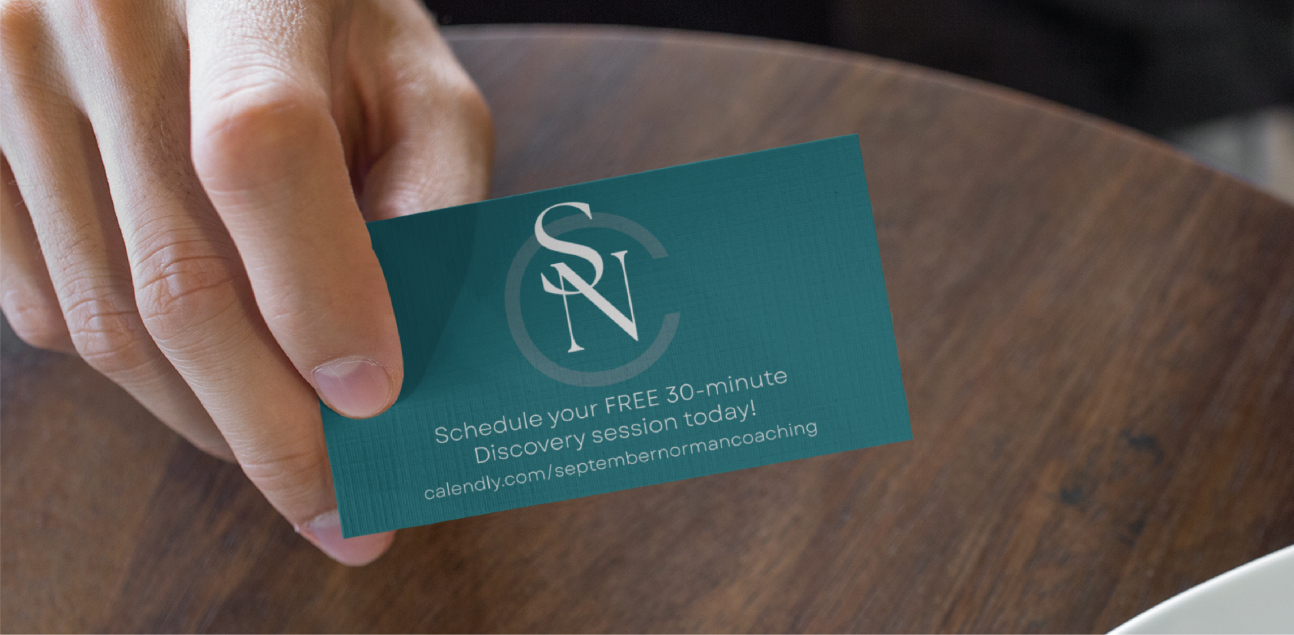 A hand holds a teal business card for a Tennessee Life Coach with the initials SN and text: Schedule your FREE 30-minute Discovery session today! calendly.com/septembenormancoaching, above a wooden table.