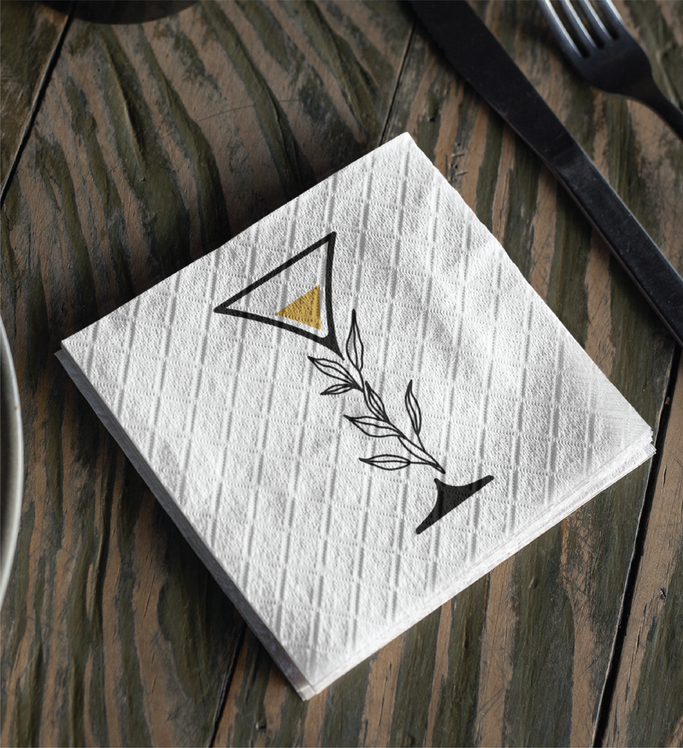A white napkin on a rustic wooden table features a black and gold martini glass with a leafy stem, showcasing a custom logo design perfect for any restaurant in Florida, next to a knife and fork.