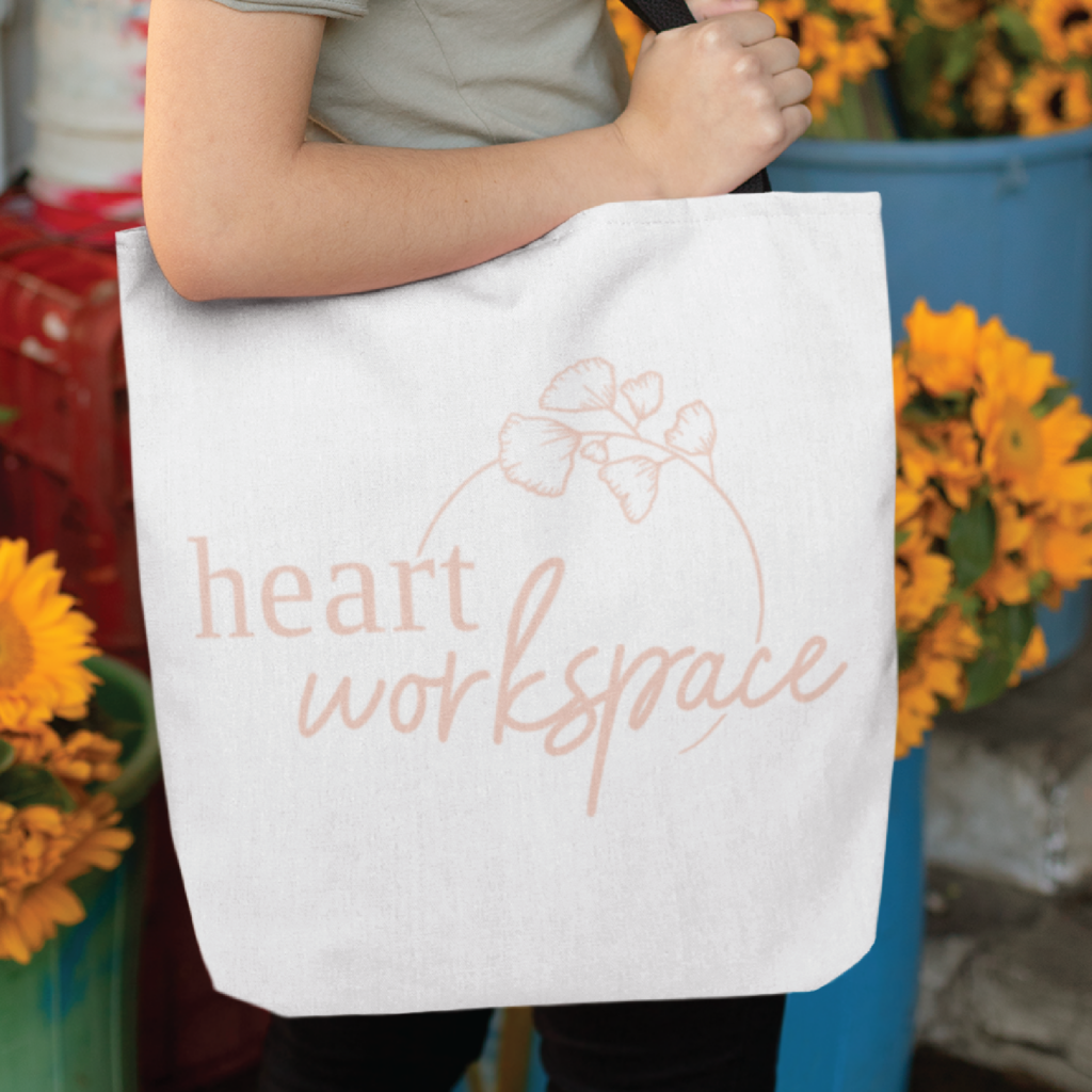 A person holds a white tote bag featuring a heart workspace logo design and delicate floral artwork, perfect for a women-focused business. Sunflowers and blue containers appear in the background.