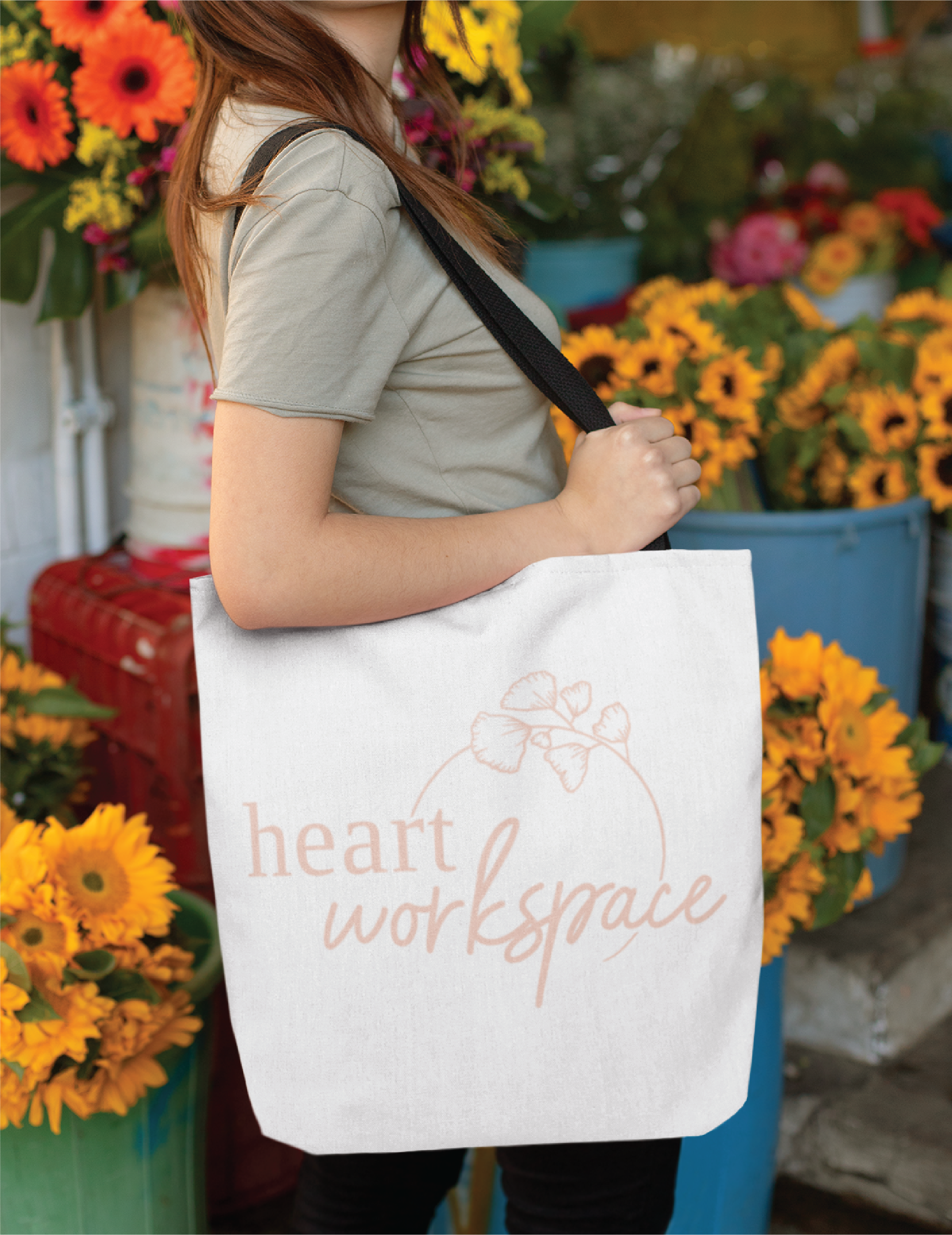 A woman stands among vibrant yellow and orange flowers in New York, holding a white tote bag with the words heart workspace and a custom logo design featuring a simple floral print.