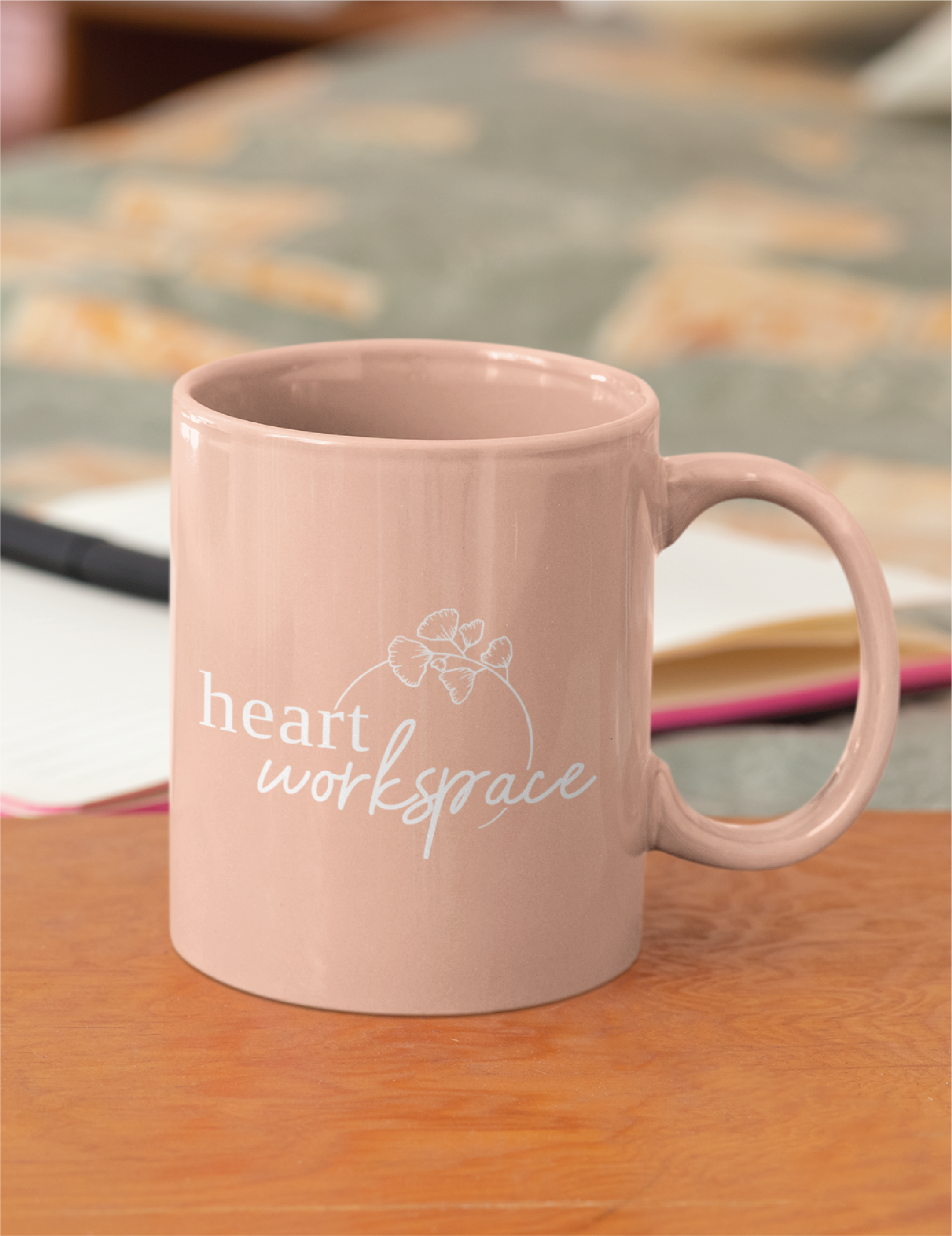 A light pink mug with the words heart workspace and a small flower graphic, featuring a custom logo design, sits on a wooden surface with a notebook and pen in the background.