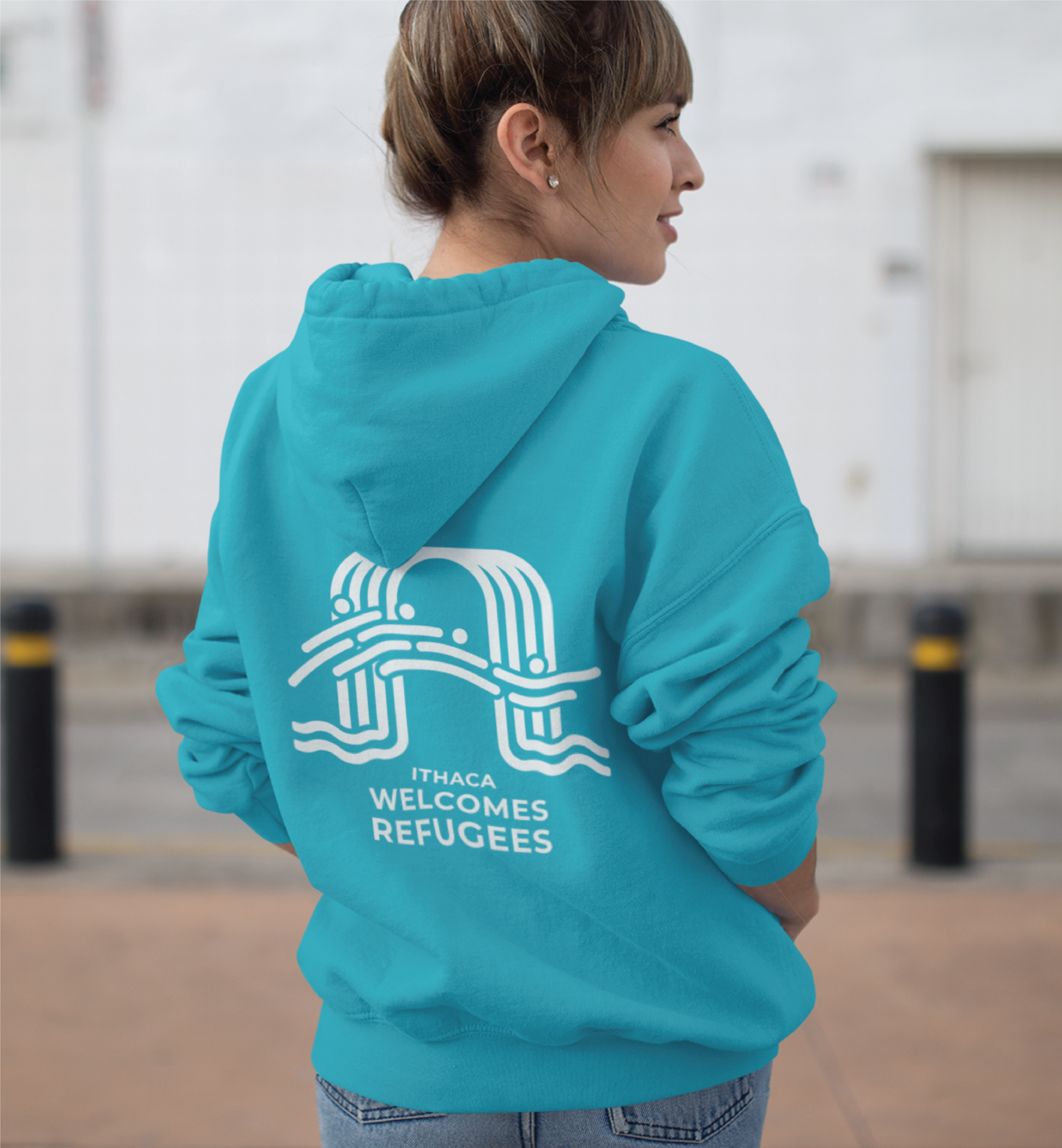 A person stands outdoors wearing a bright blue hoodie with a white graphic and the text ITHACA WELCOMES REFUGEES on the back, showcasing a New custom logo design for the nonprofit organization. The background features a white building and black post barriers.