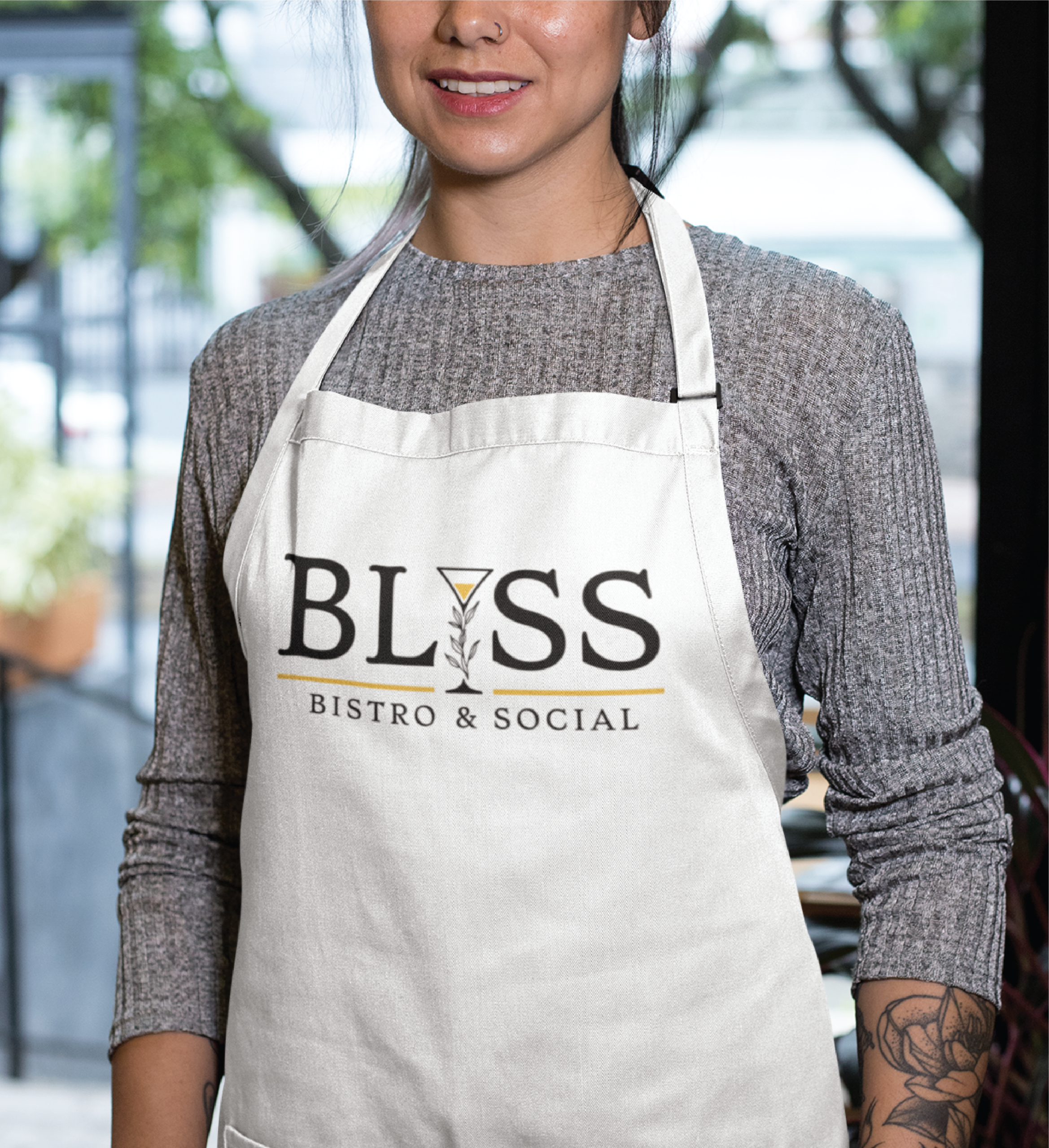 A woman wearing a light gray ribbed sweater and a white apron with the custom logo design BLISS Bistro & Social stands indoors, smiling slightly. Greenery and large windows blur in the background, evoking a welcoming Florida restaurant vibe.