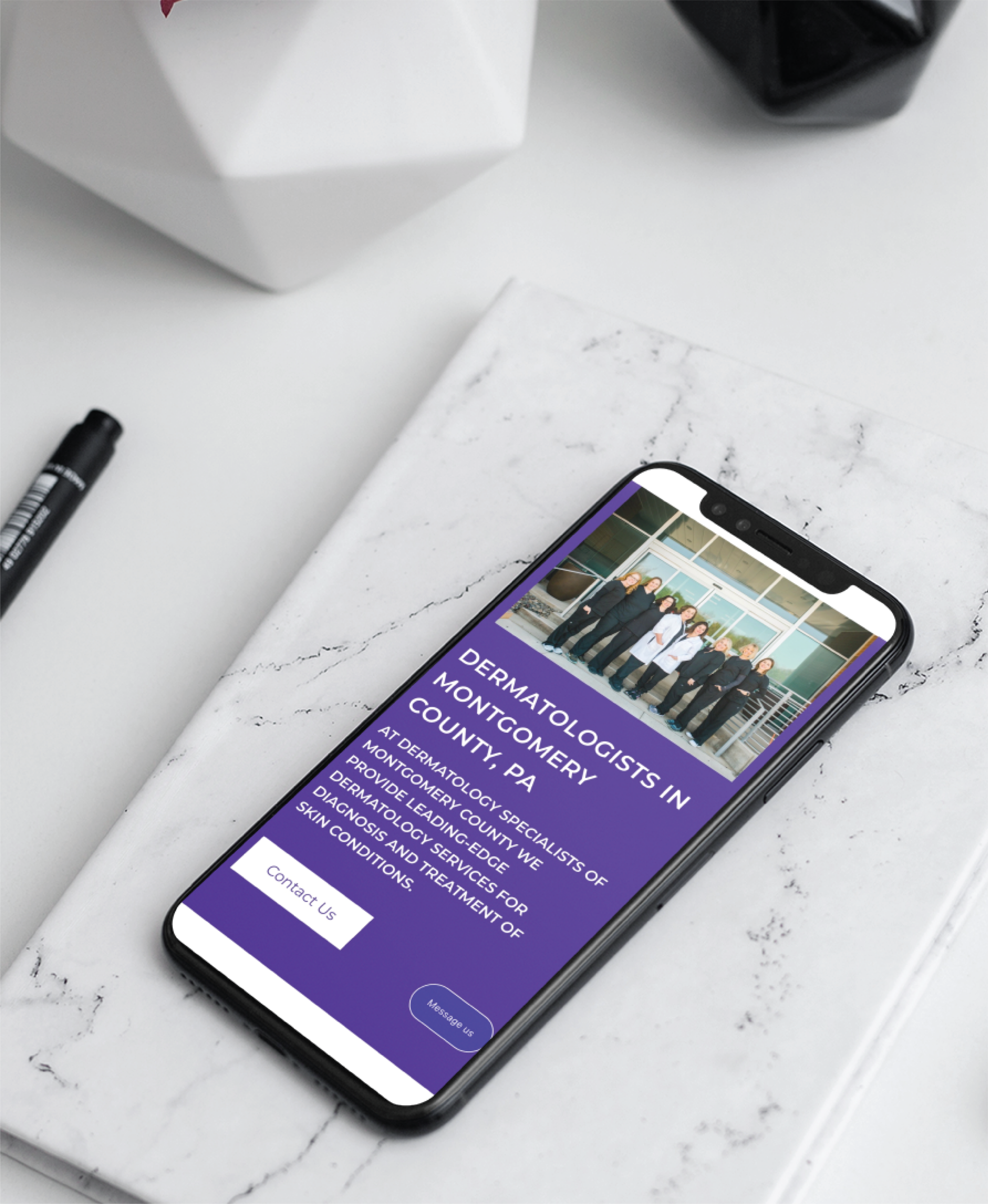 A smartphone on a marble surface displays a medical practice website for dermatologists in Montgomery County, Pennsylvania, featuring a group photo, description, and Contact Us button. A pen and geometric decor are nearby.